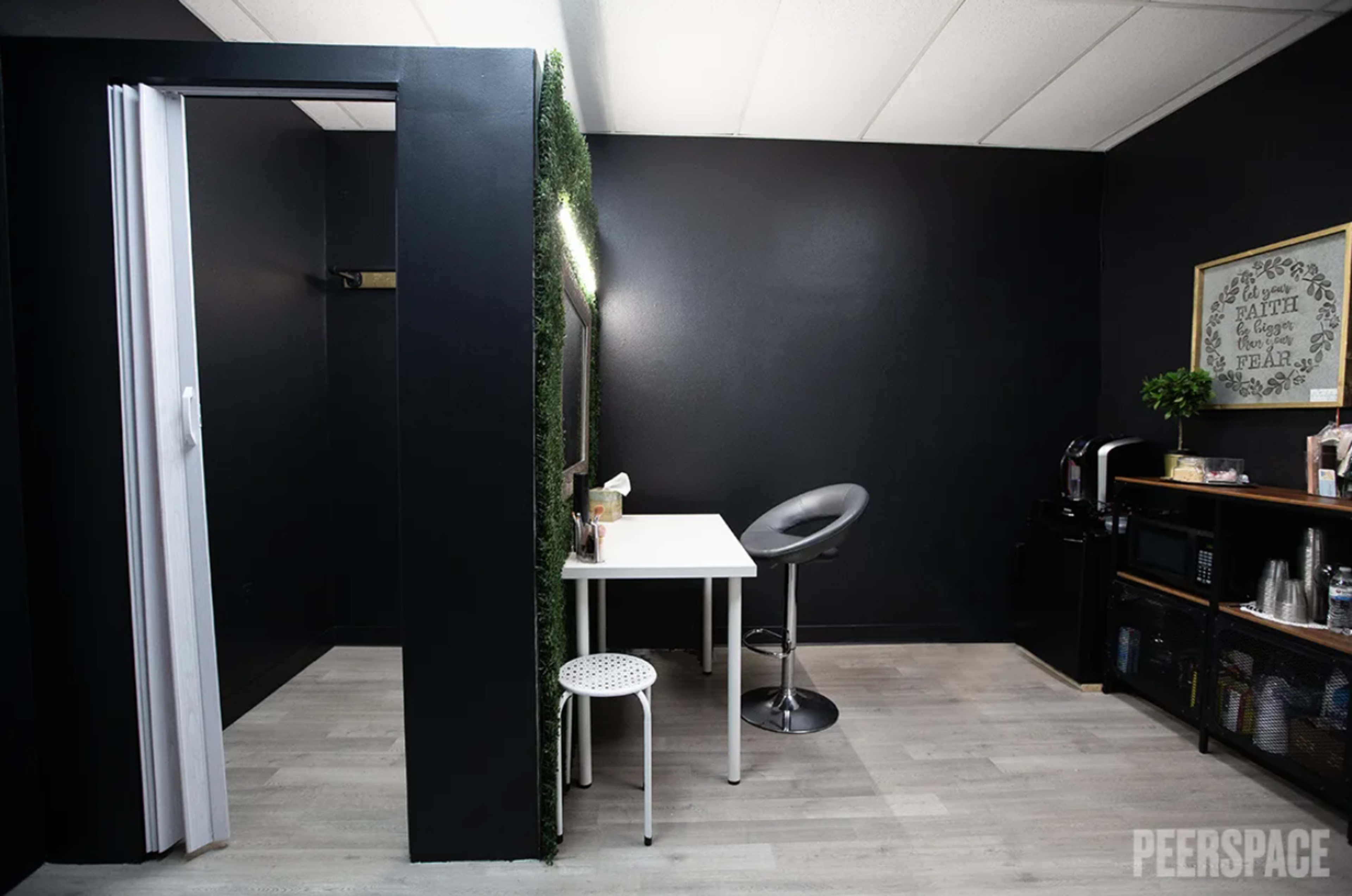 The image shows a small room with black walls, featuring a white table with a bar stool, a small chair, and a coffee station on the side.