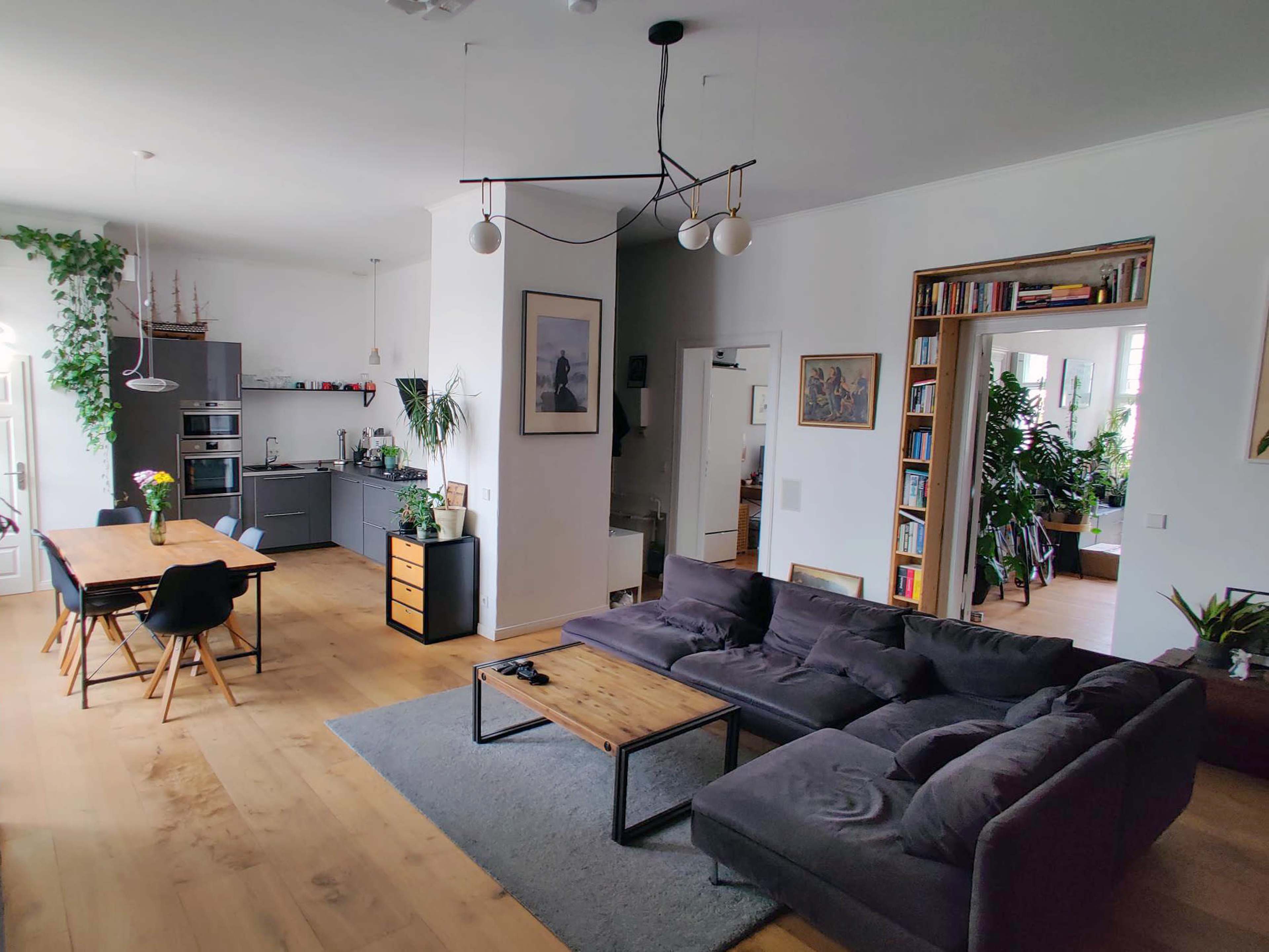 A spacious living area with a gray sectional sofa, a wooden coffee table, a dining table with chairs, and a modern kitchen in the background.