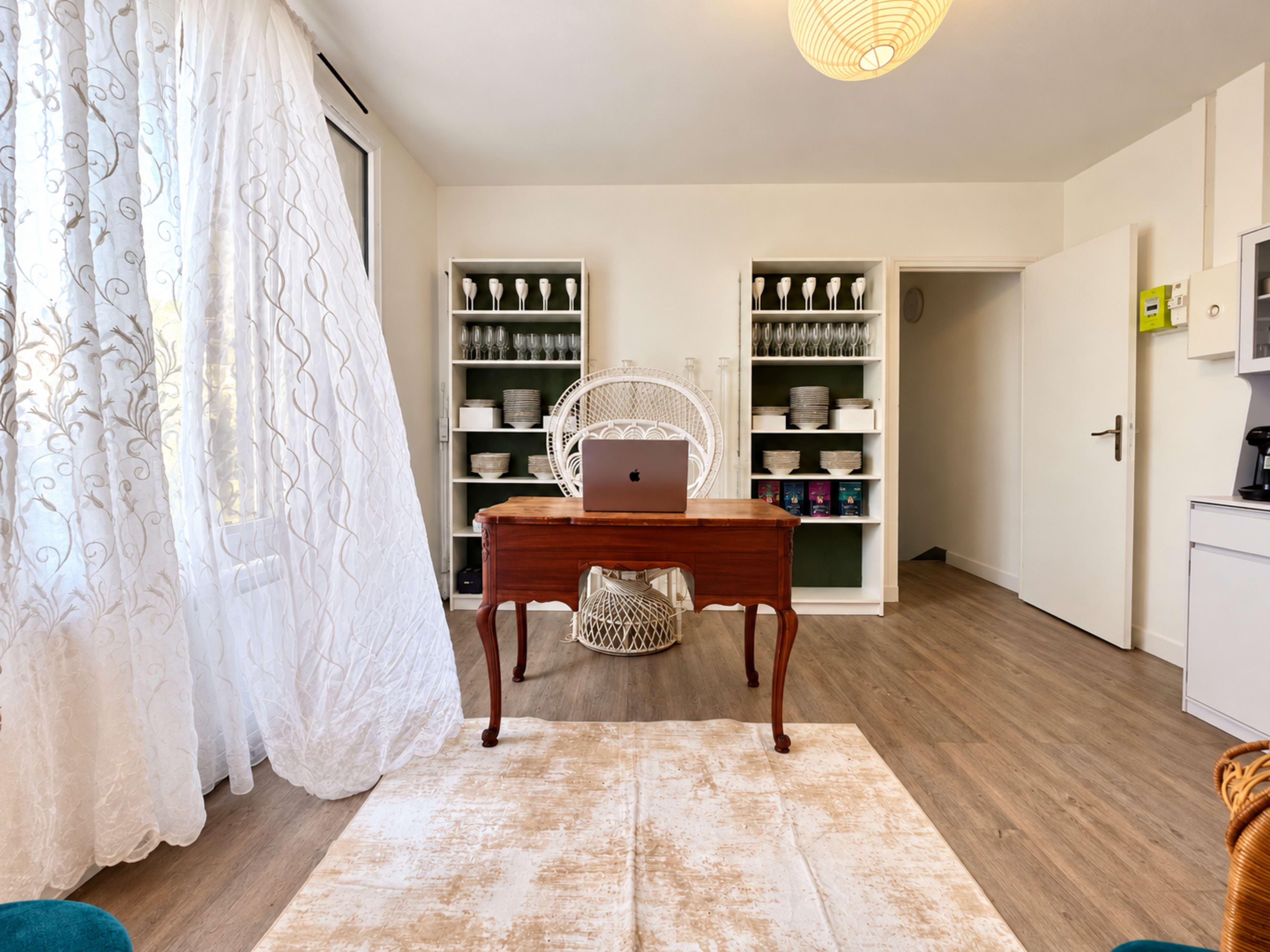 The room features a wooden desk with a laptop, surrounded by shelves filled with dishware and a large window draped with sheer curtains.