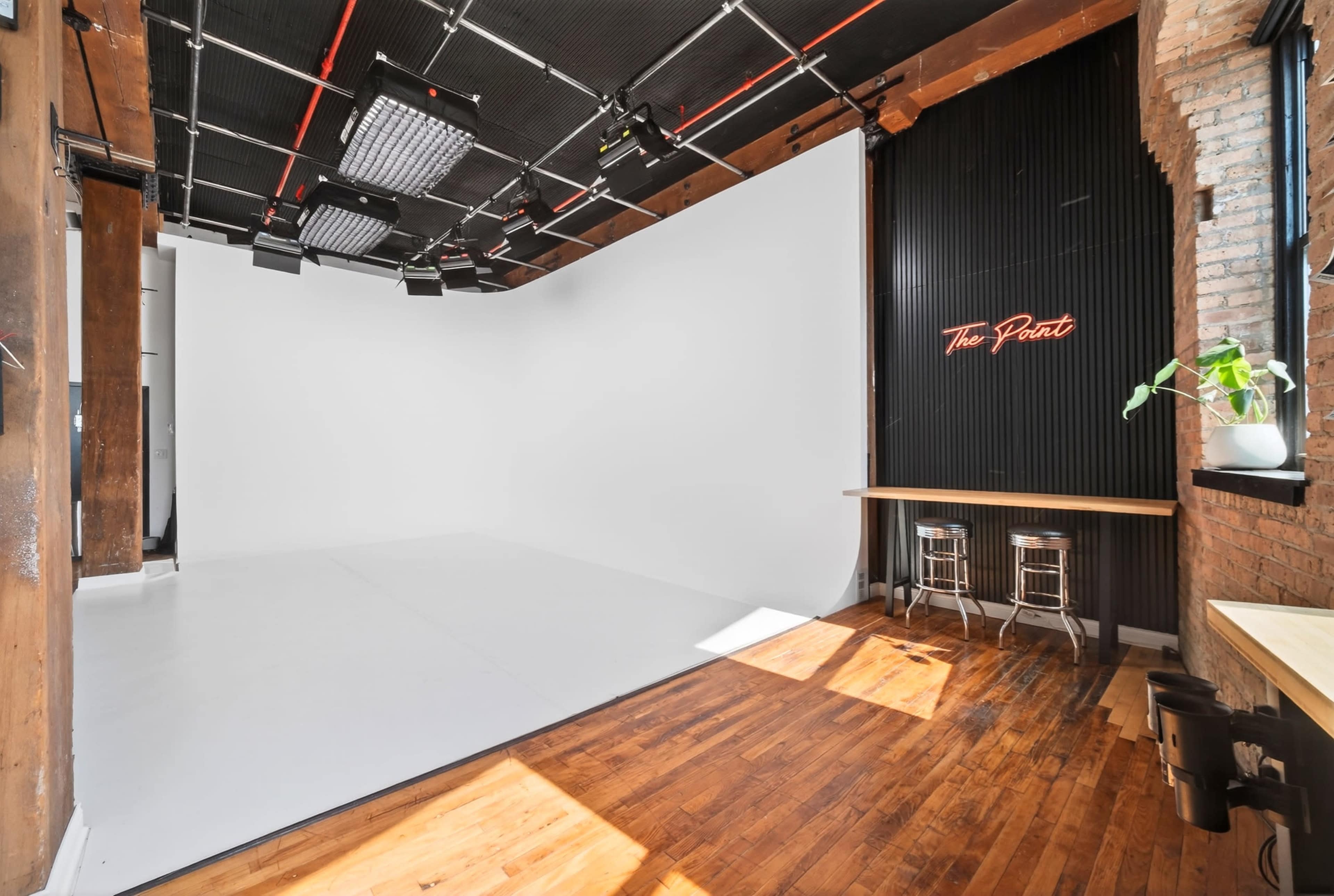 The image shows a photography studio with a large white backdrop, wooden flooring, and a bar area with two stools against a textured wall featuring neon text.