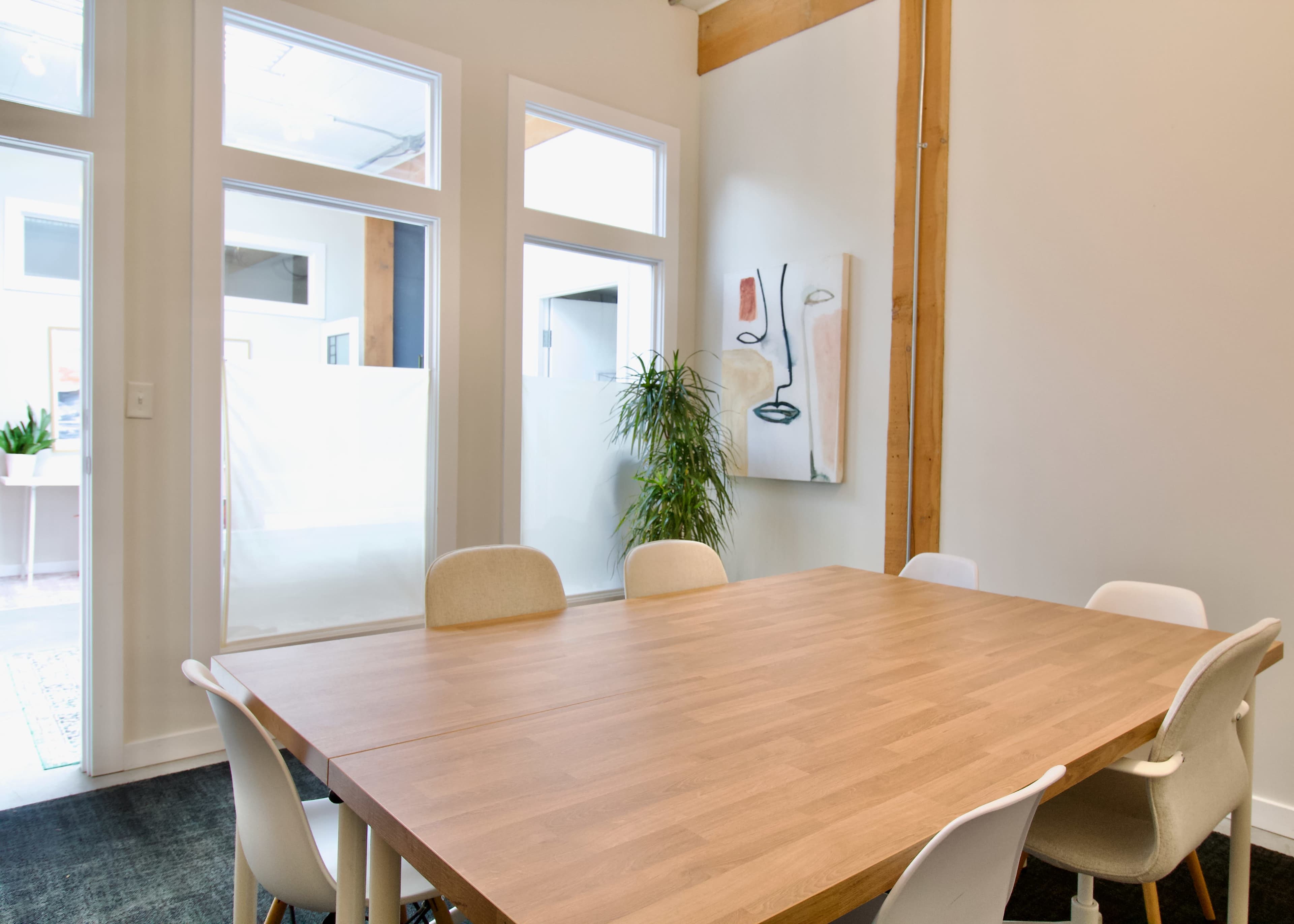 The image shows a modern conference room with a large wooden table and white chairs, featuring a plant and artwork on the wall.