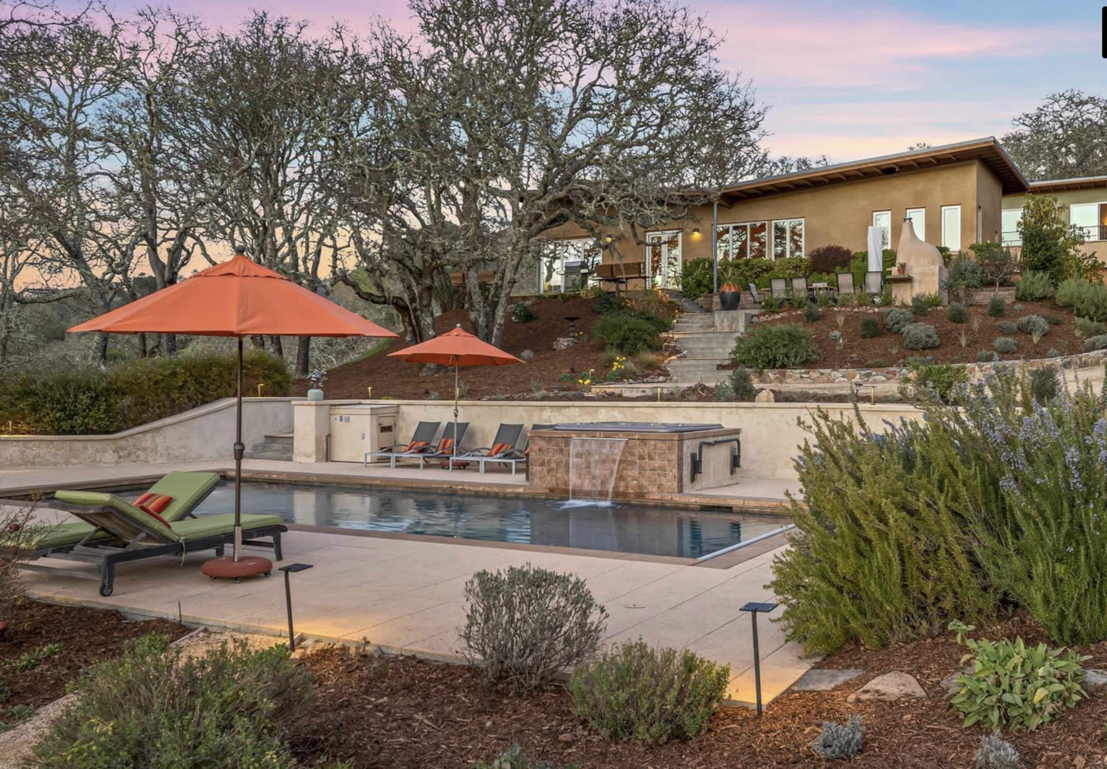 A landscaped backyard features a pool with a spa, surrounded by lounge chairs and umbrellas, alongside a modern house set against a backdrop of trees.