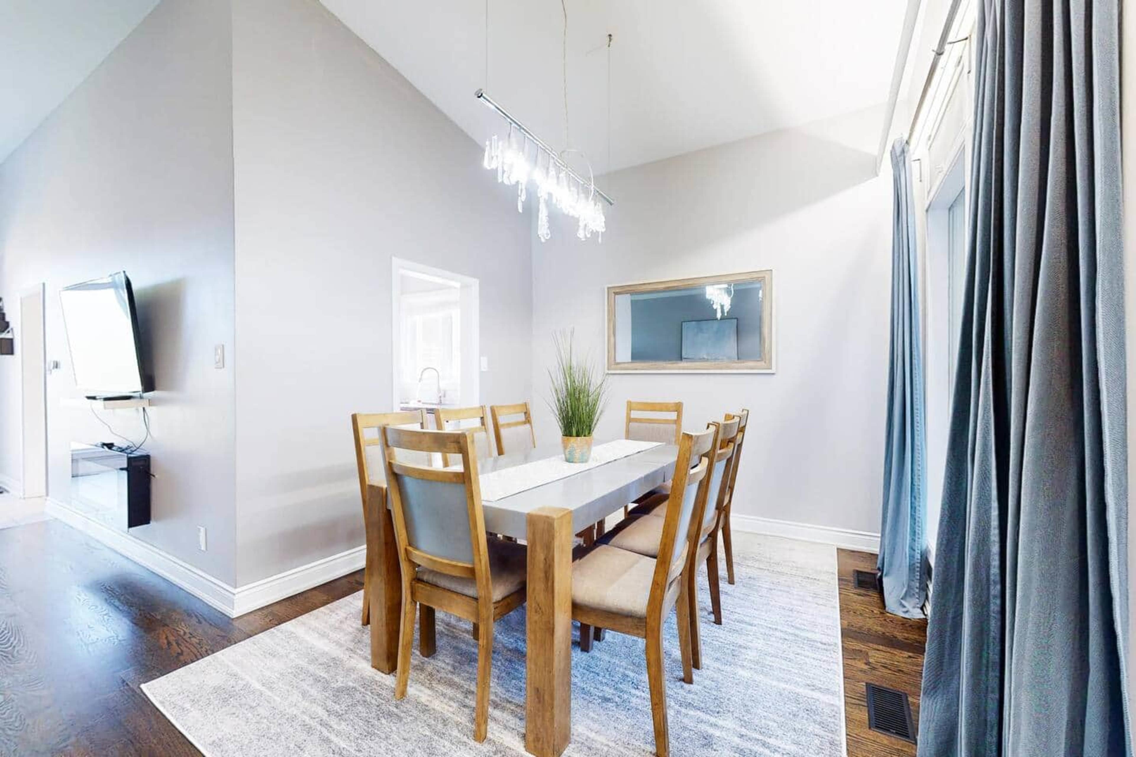 The image shows a well-lit dining area featuring a rectangular table surrounded by several wooden chairs, a chandelier overhead, and a mirror on the wall.