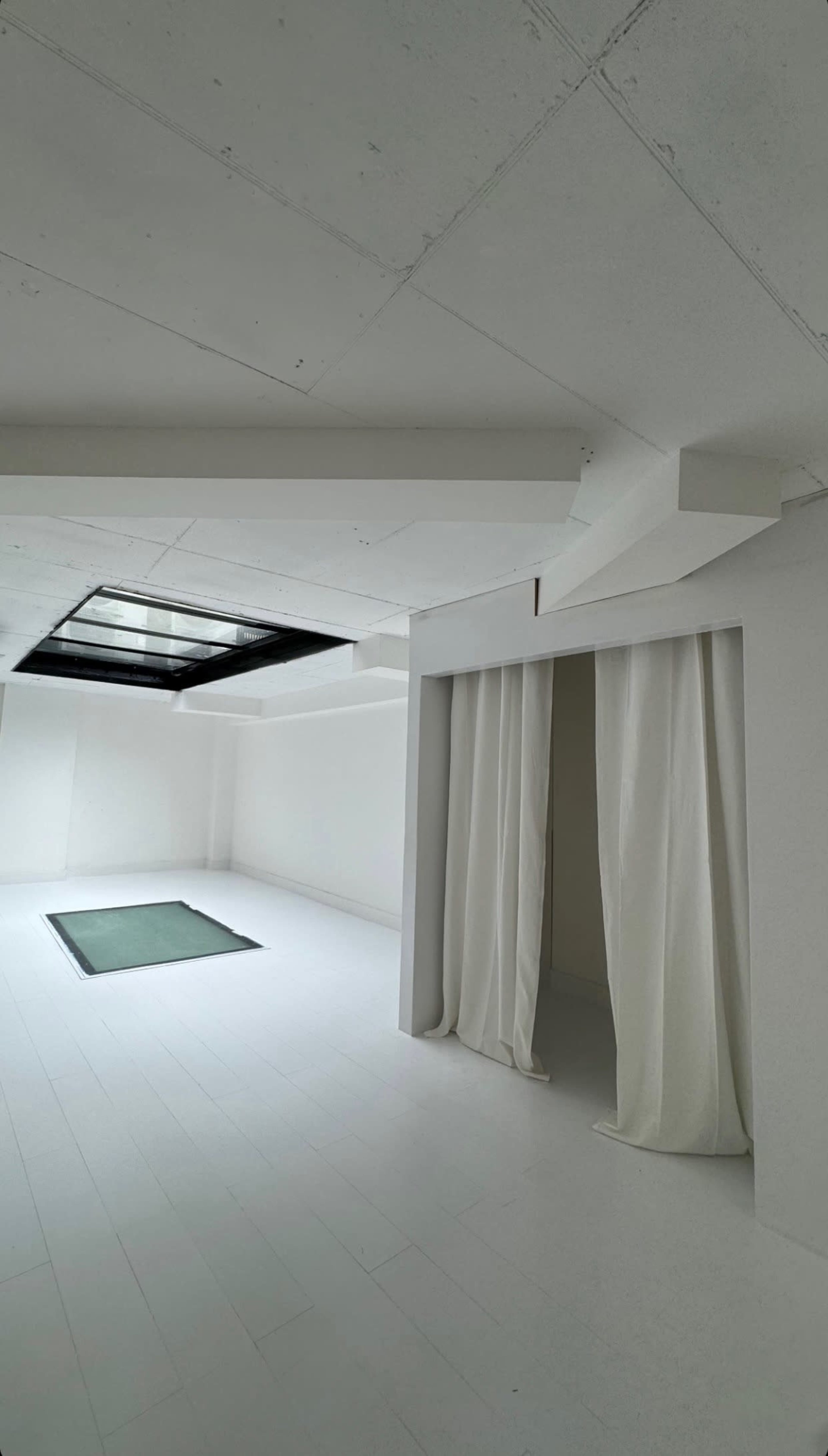 A minimalistic interior space with white walls, a green rectangular mat on the floor, and a pair of white curtains partially drawn to one side.