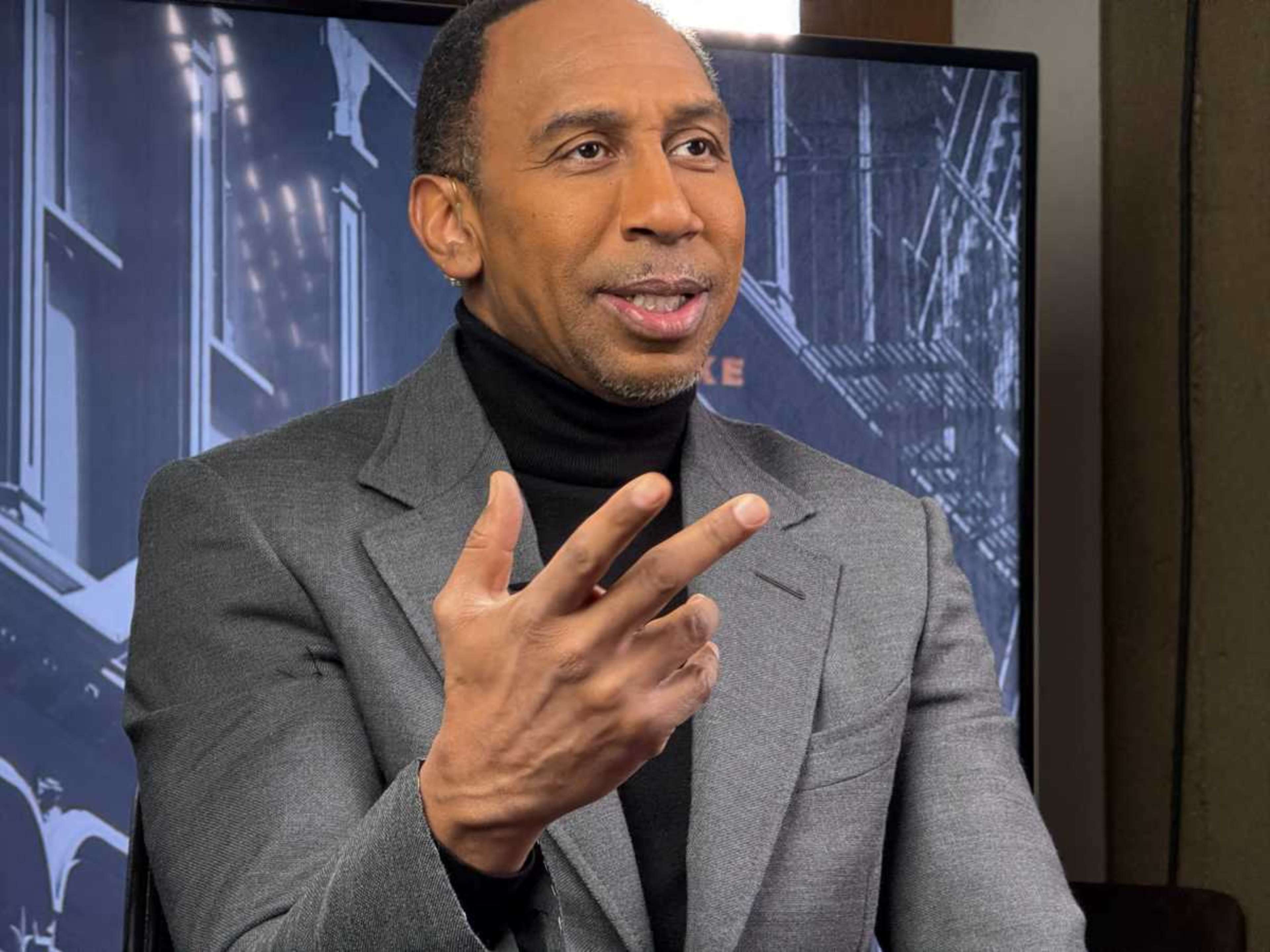 A man in a gray suit jacket gestures while speaking, with a blurred background that includes a screen displaying text.