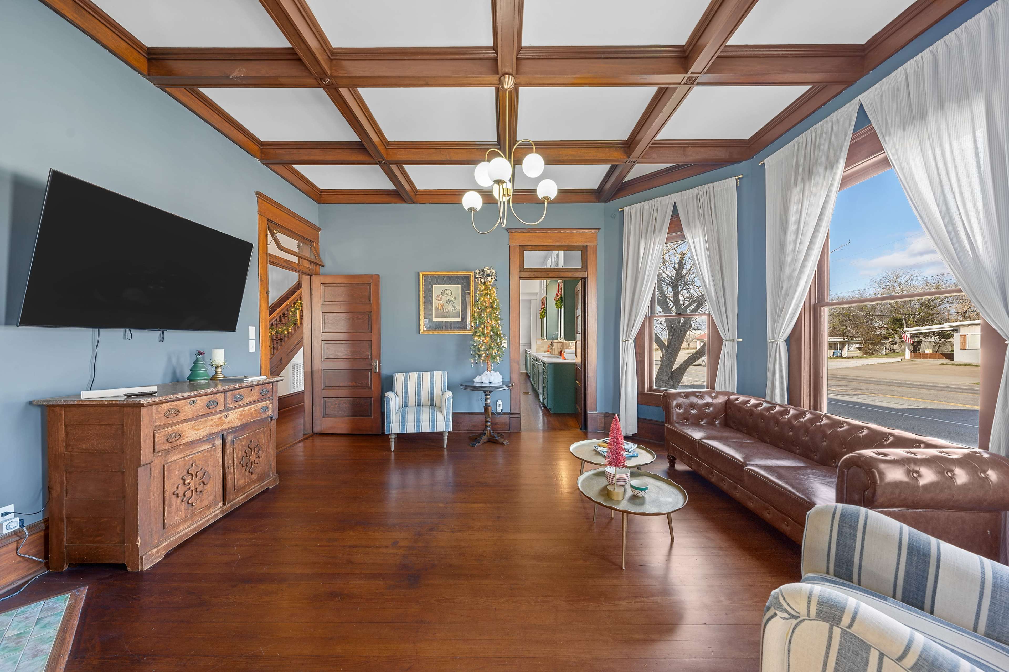 A spacious living area with a coffered ceiling, a leather sofa set, a wooden cabinet, and large windows allowing natural light to enter.