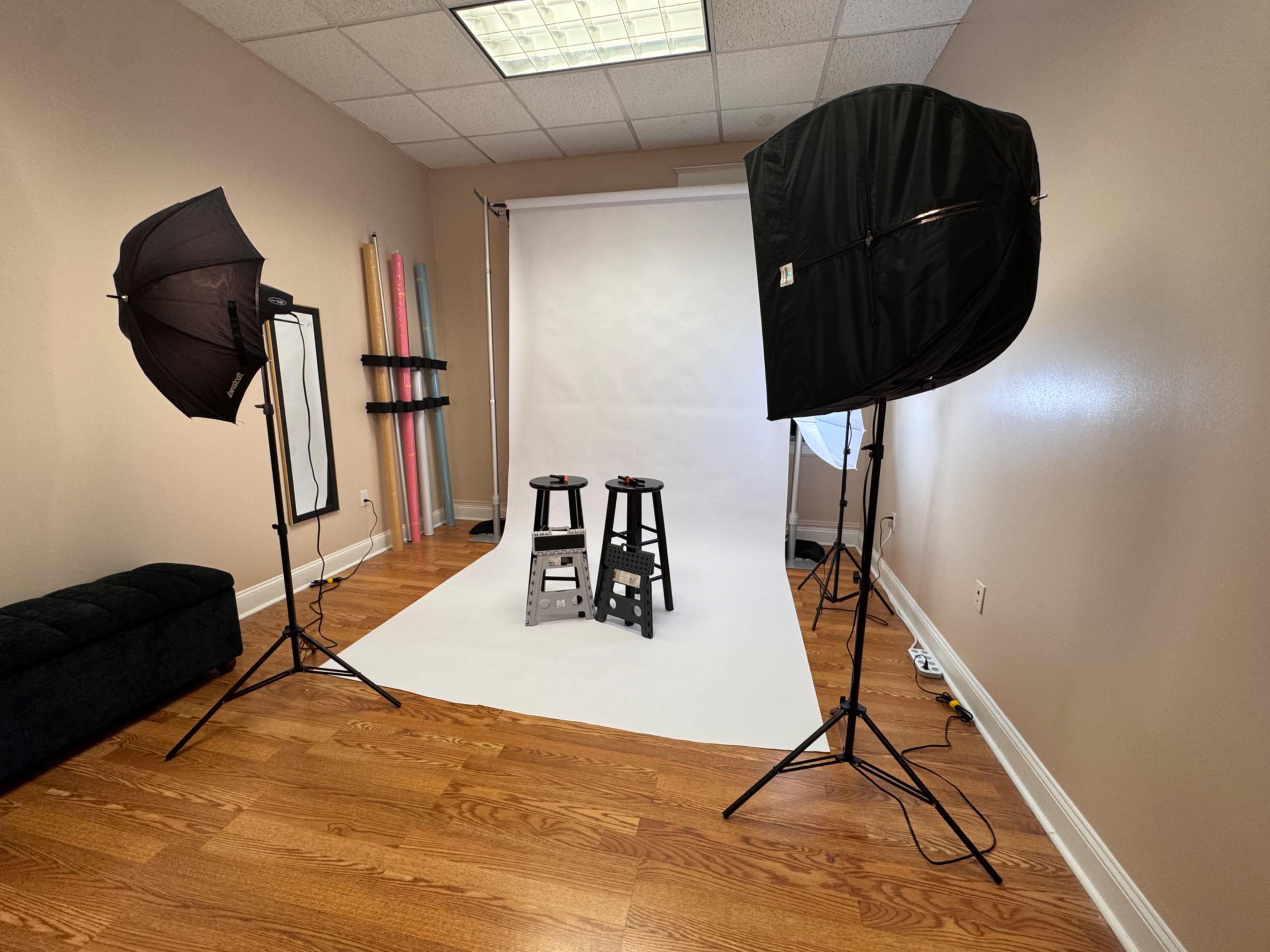 The image shows a photo studio with two black stools positioned on a white backdrop, flanked by two softbox light setups.