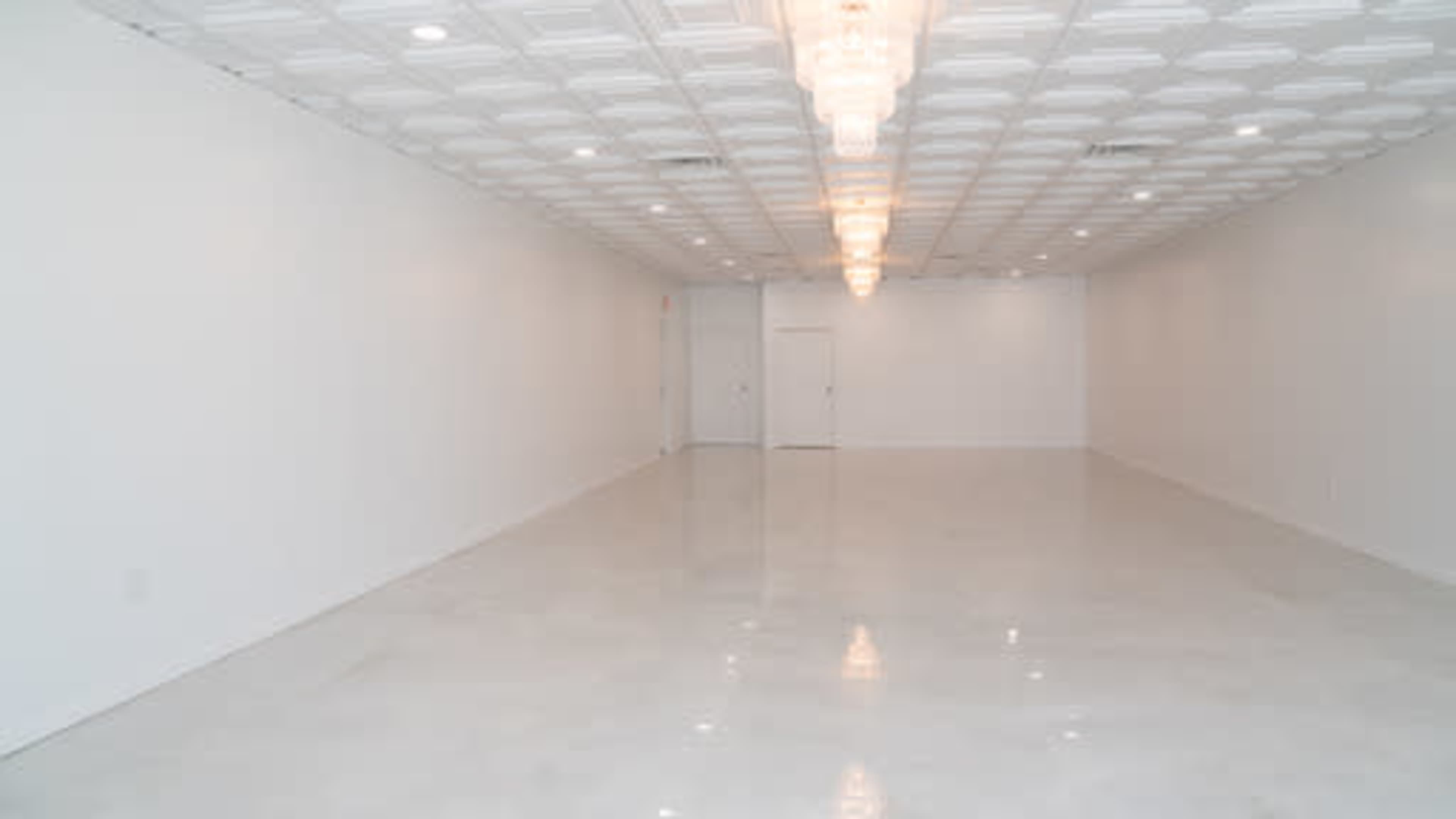 The image shows a spacious, empty room with white walls and a shiny floor, illuminated by a chandelier overhead.