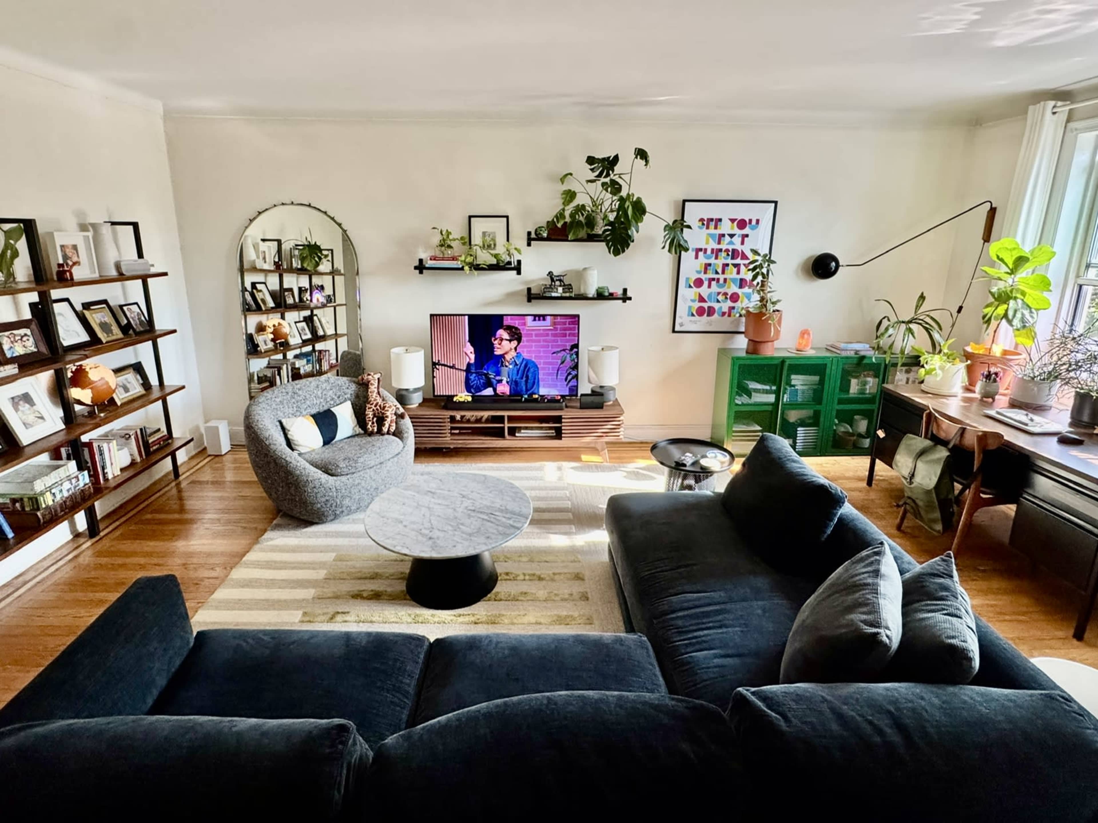The image shows a cozy living room with a large dark sectional sofa, a round coffee table, a television displaying a program, and several plants arranged on shelves and tables around the room.
