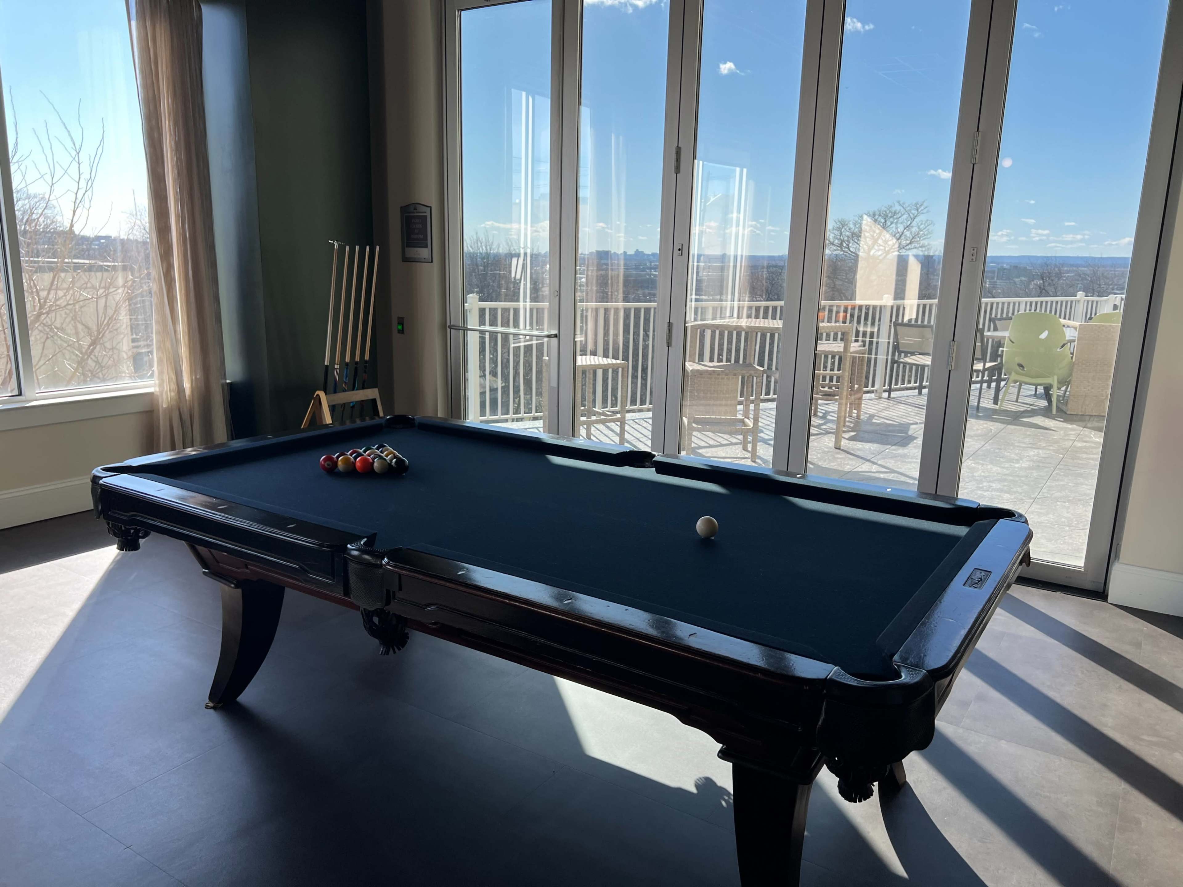 A black pool table sits in a brightly lit room with large windows overlooking an outdoor terrace.