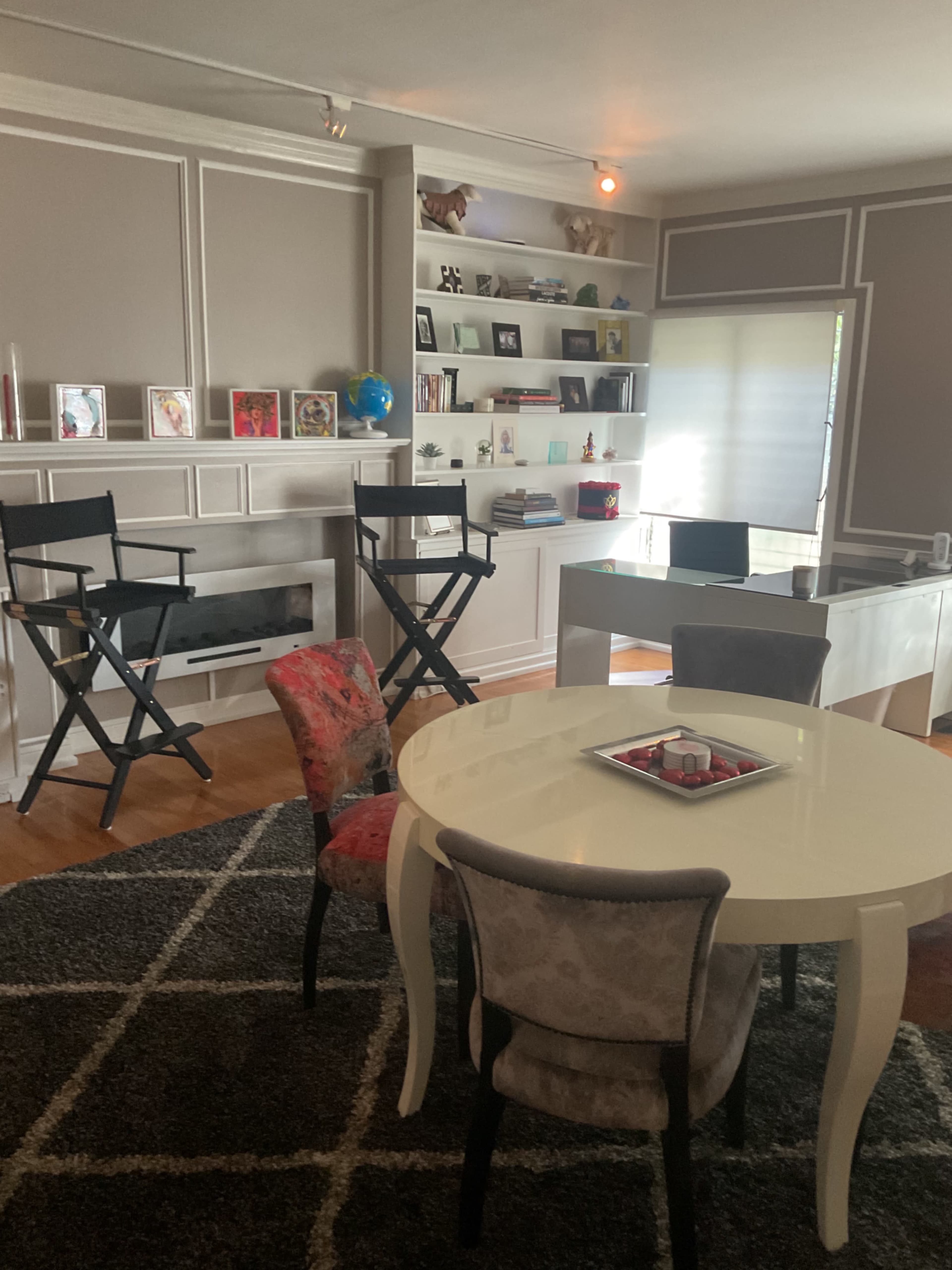 The image shows a modern office space featuring a round table, a desk, two director's chairs, and a bookshelf filled with various items.