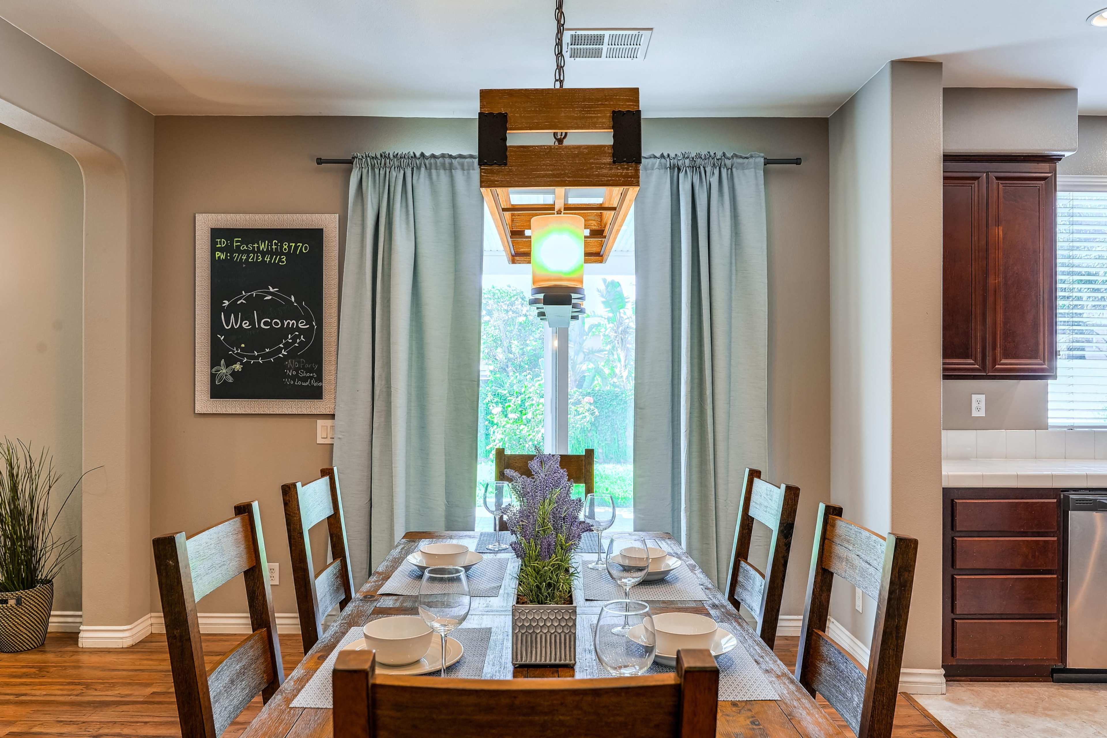 A wooden dining table is set for a meal, with plates arranged on it, positioned near a window with light green curtains and a chalkboard displaying a welcome message.