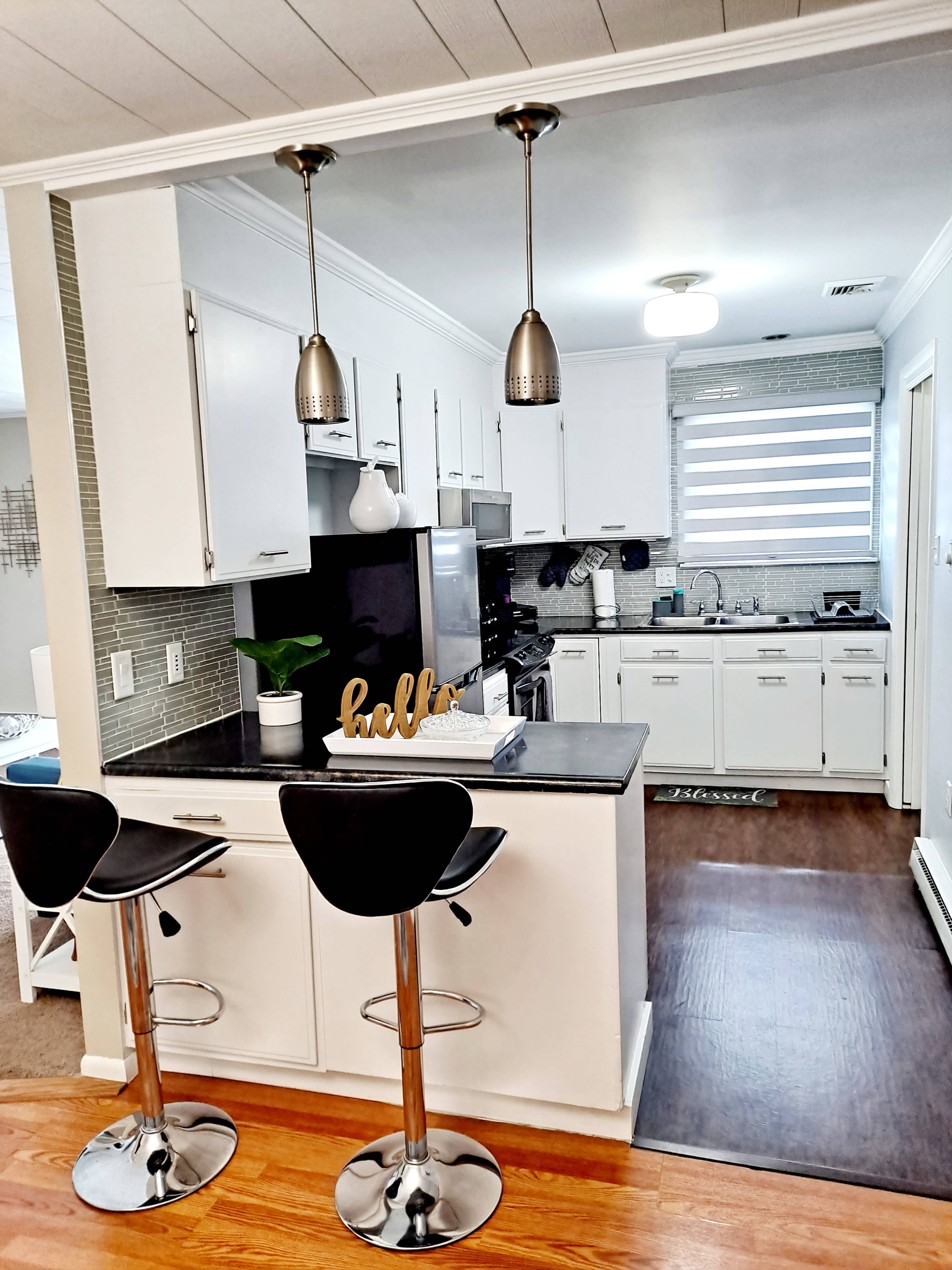 The image shows a modern kitchen with a black countertop, white cabinets, two bar stools, and pendant lighting.