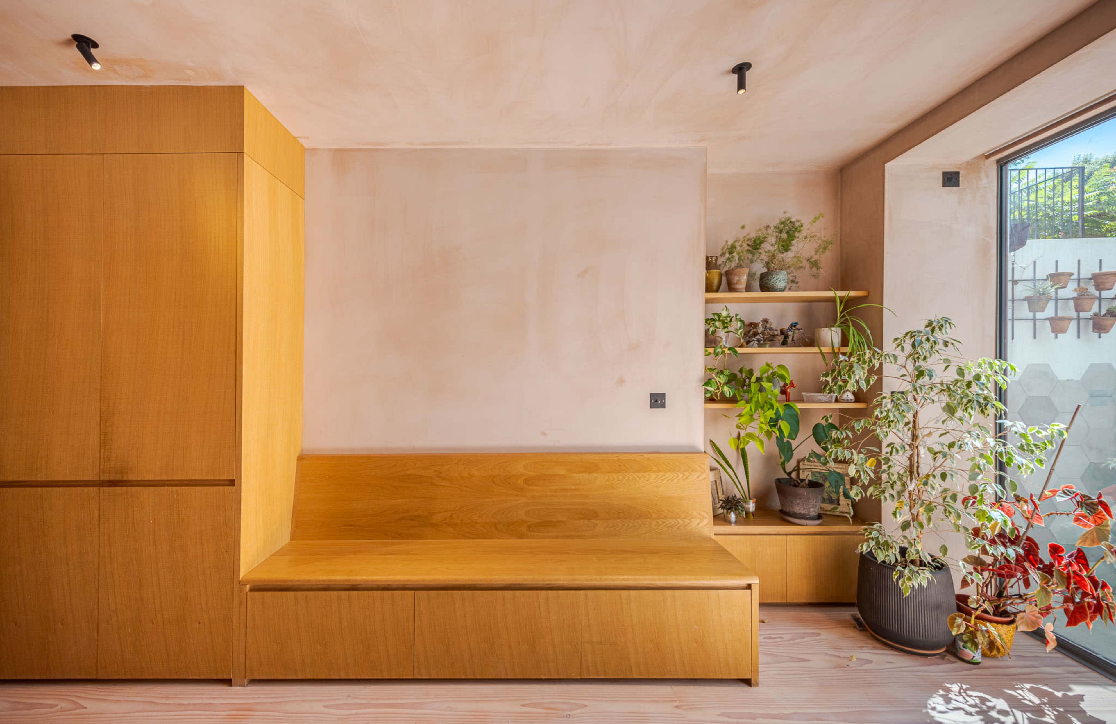 The image shows a minimalist interior with wooden furniture, a built-in bench, and plants near a window.