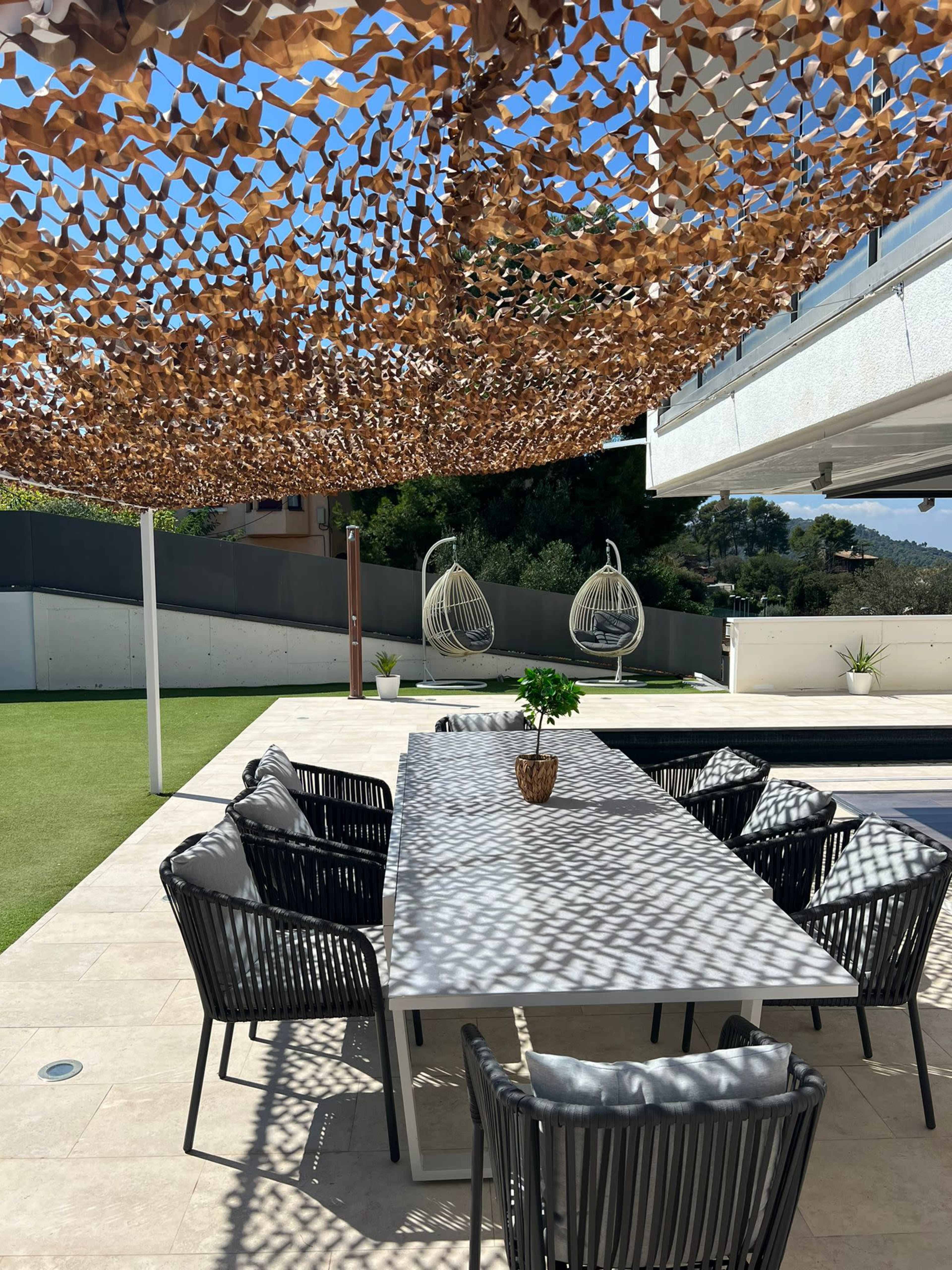 A modern outdoor dining area features a long table surrounded by black chairs, with a decorative overhead shade and two hanging chairs in the background.