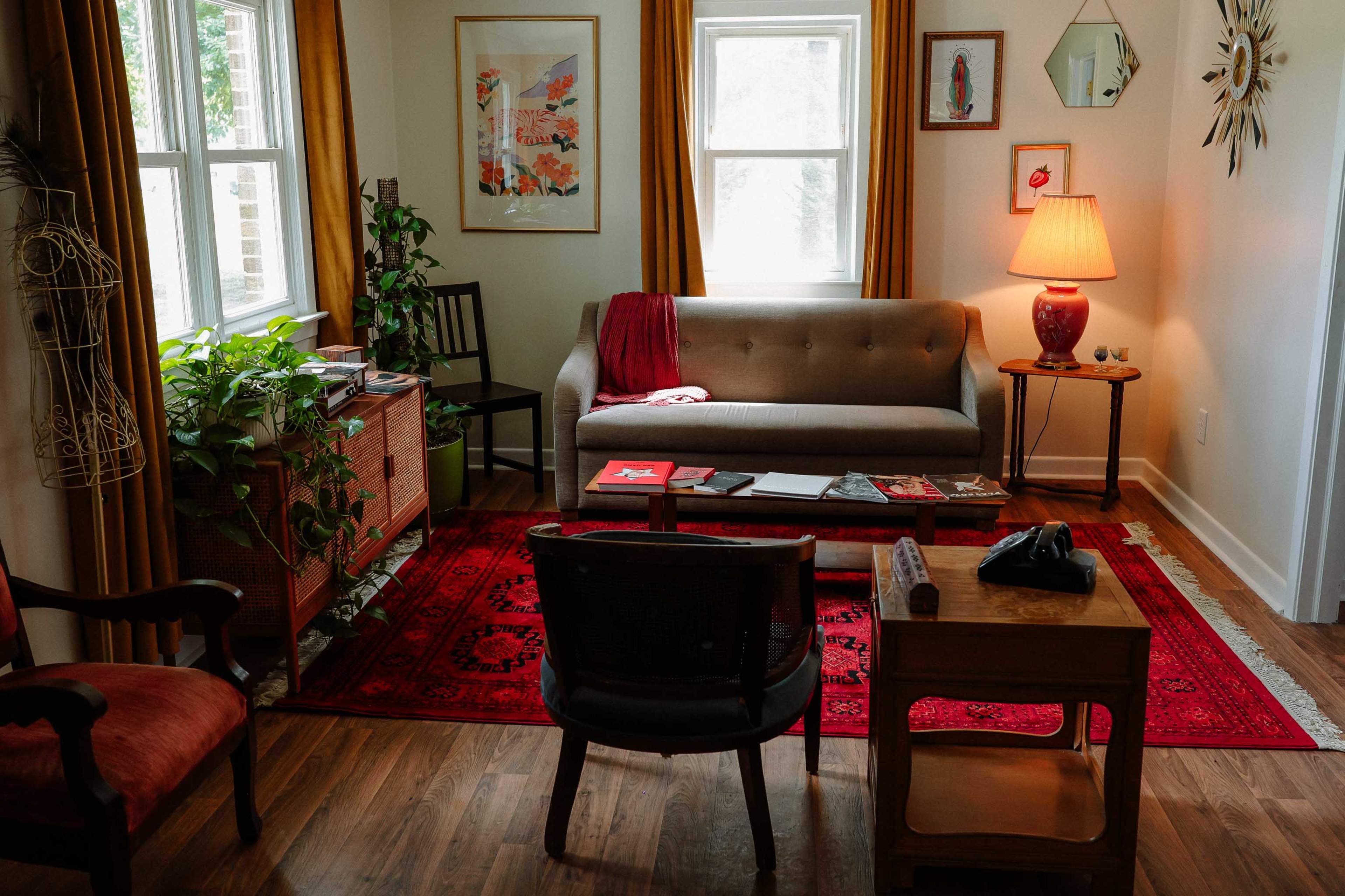 The image shows a cozy living room featuring a gray sofa, a red area rug, and various pieces of furniture including chairs and a side table, along with decorative plants and wall art.