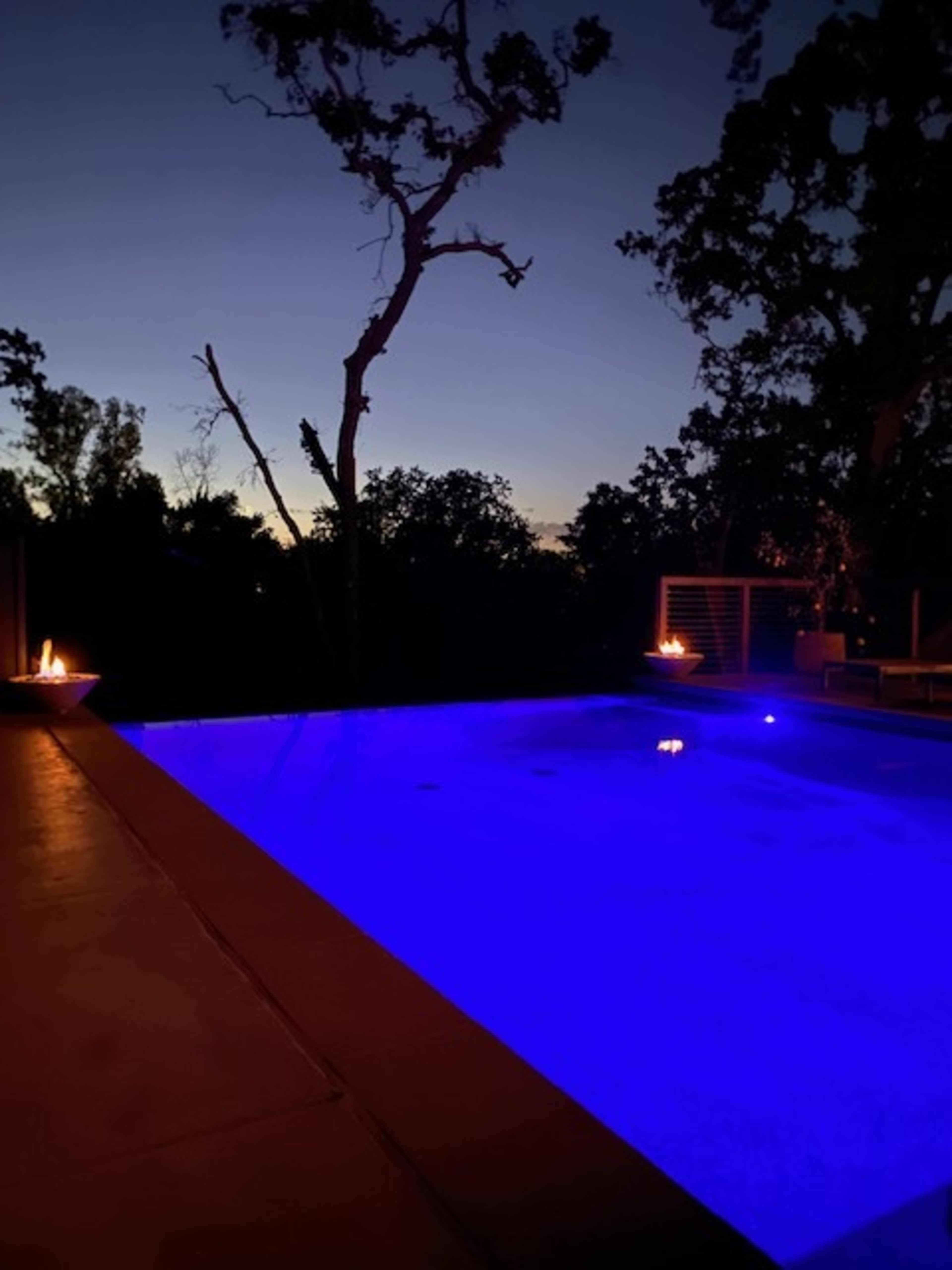 A lit swimming pool at dusk, surrounded by trees and under a fading blue sky.