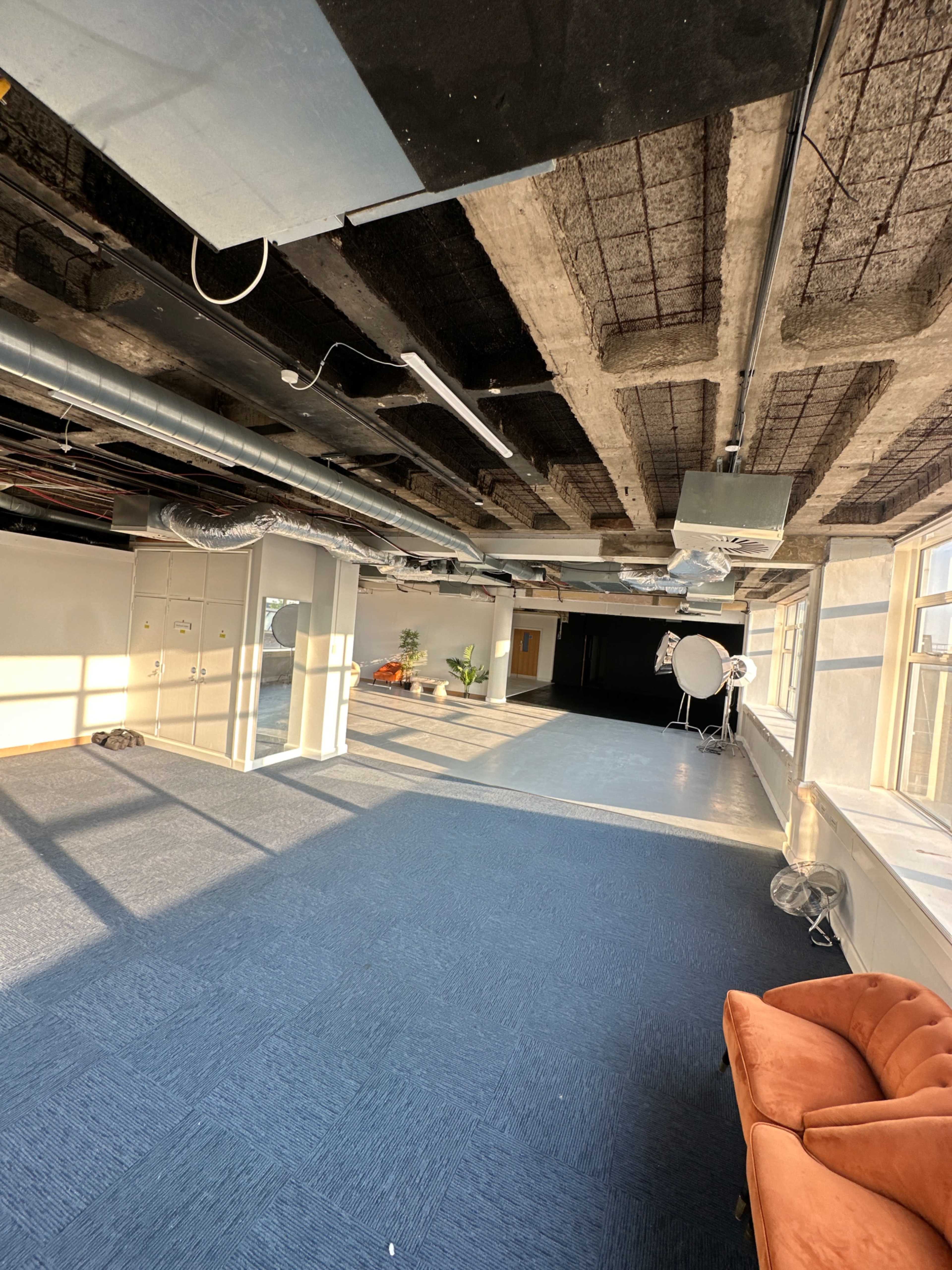 The image shows an empty room with blue carpeting, exposed ceilings, and large windows allowing sunlight to stream in.