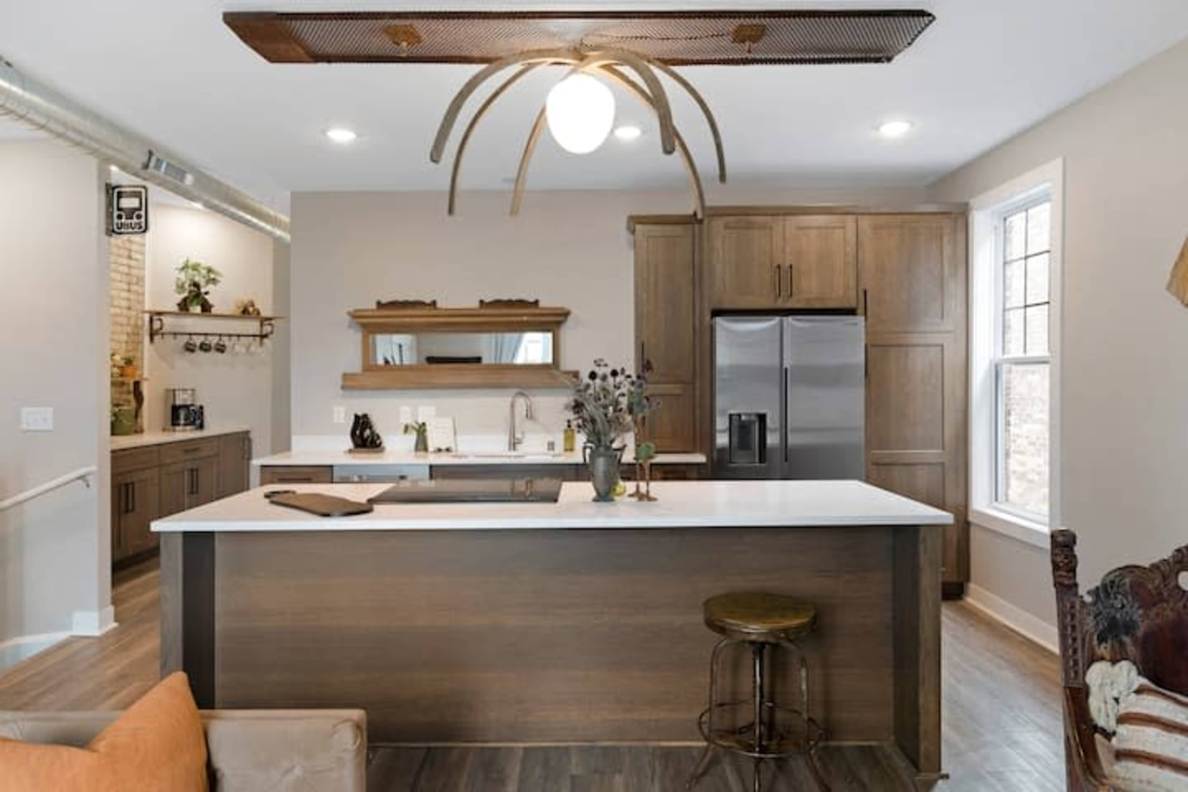 The image shows a modern kitchen with wooden cabinets, a large island featuring a white countertop, and a stainless steel refrigerator.