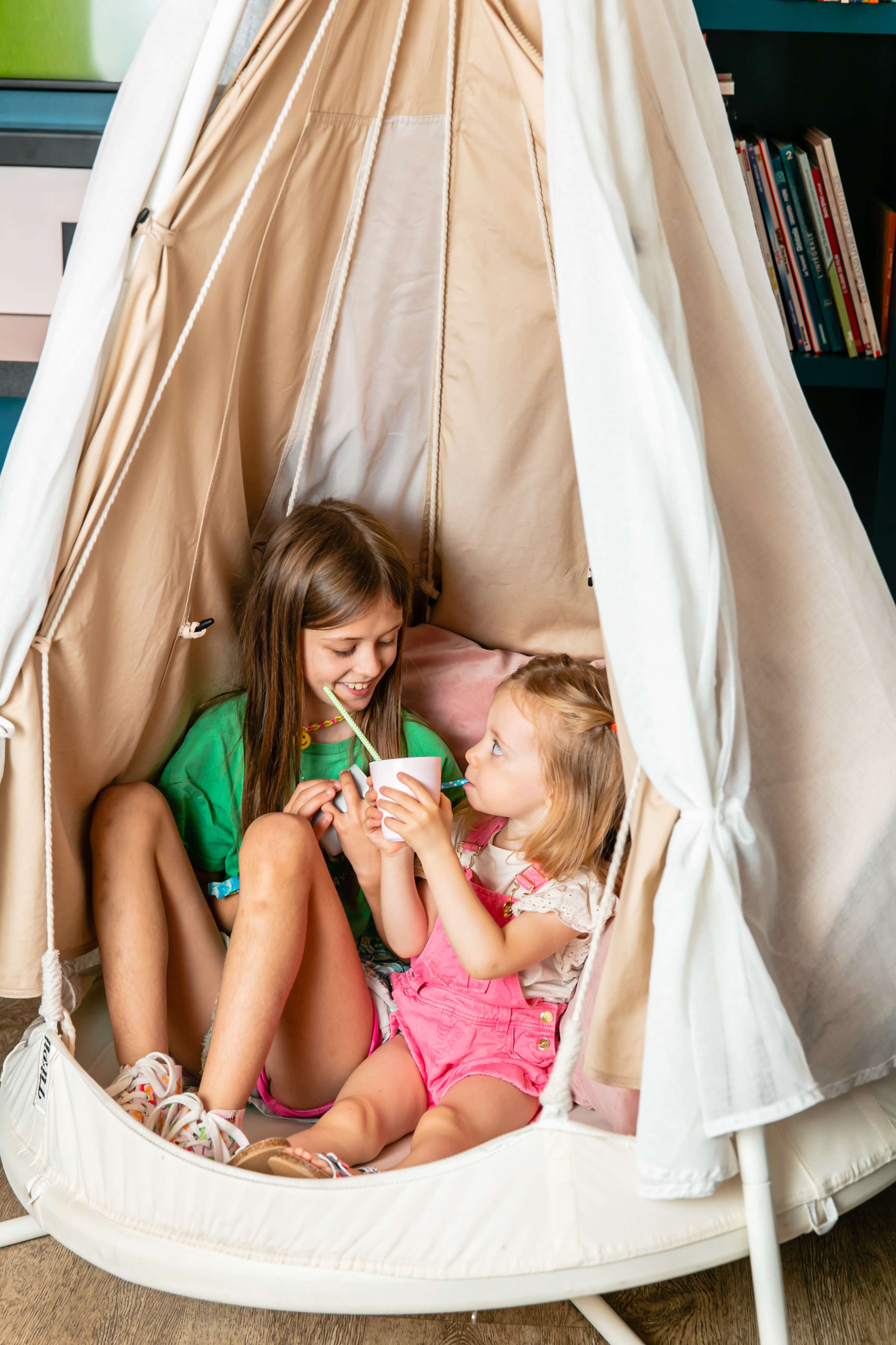 Two young girls sit inside a beige teepee, smiling and sharing a small cup.