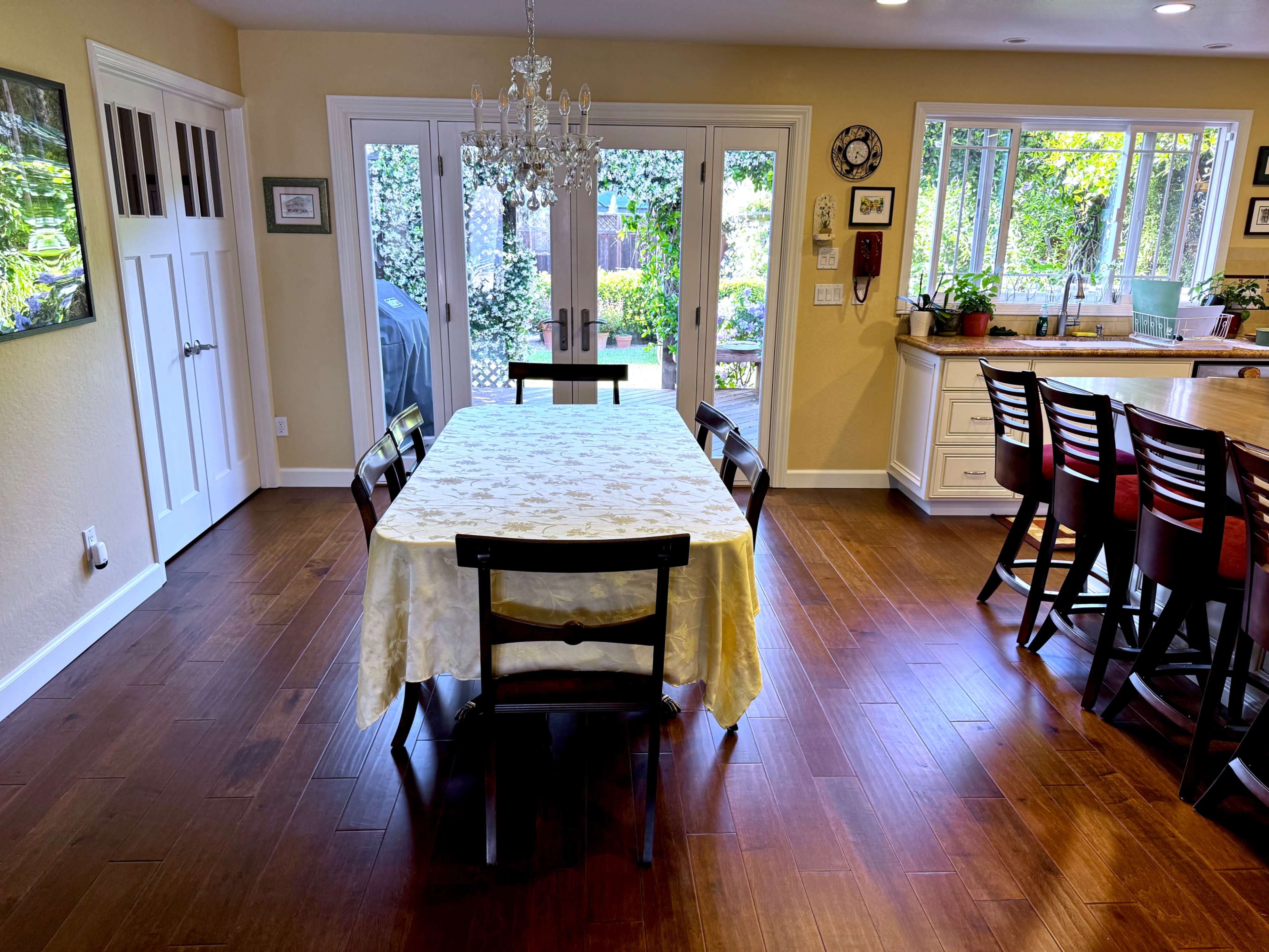 A dining area features a wooden table with a light-colored tablecloth surrounded by chairs, illuminated by a chandelier, with sliding glass doors leading to a garden view.