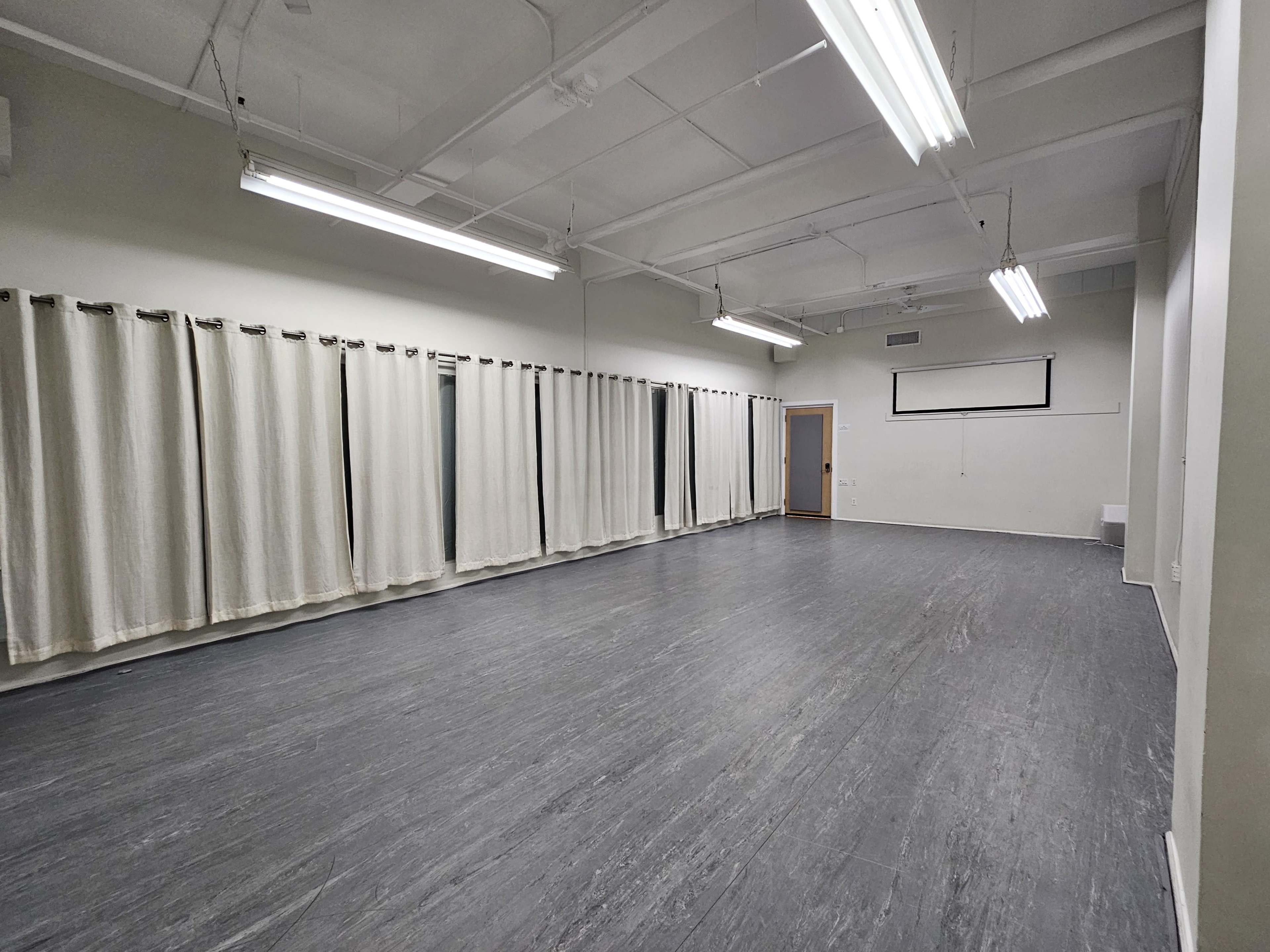 An empty, well-lit room with gray flooring and cream-colored curtains covering the windows.