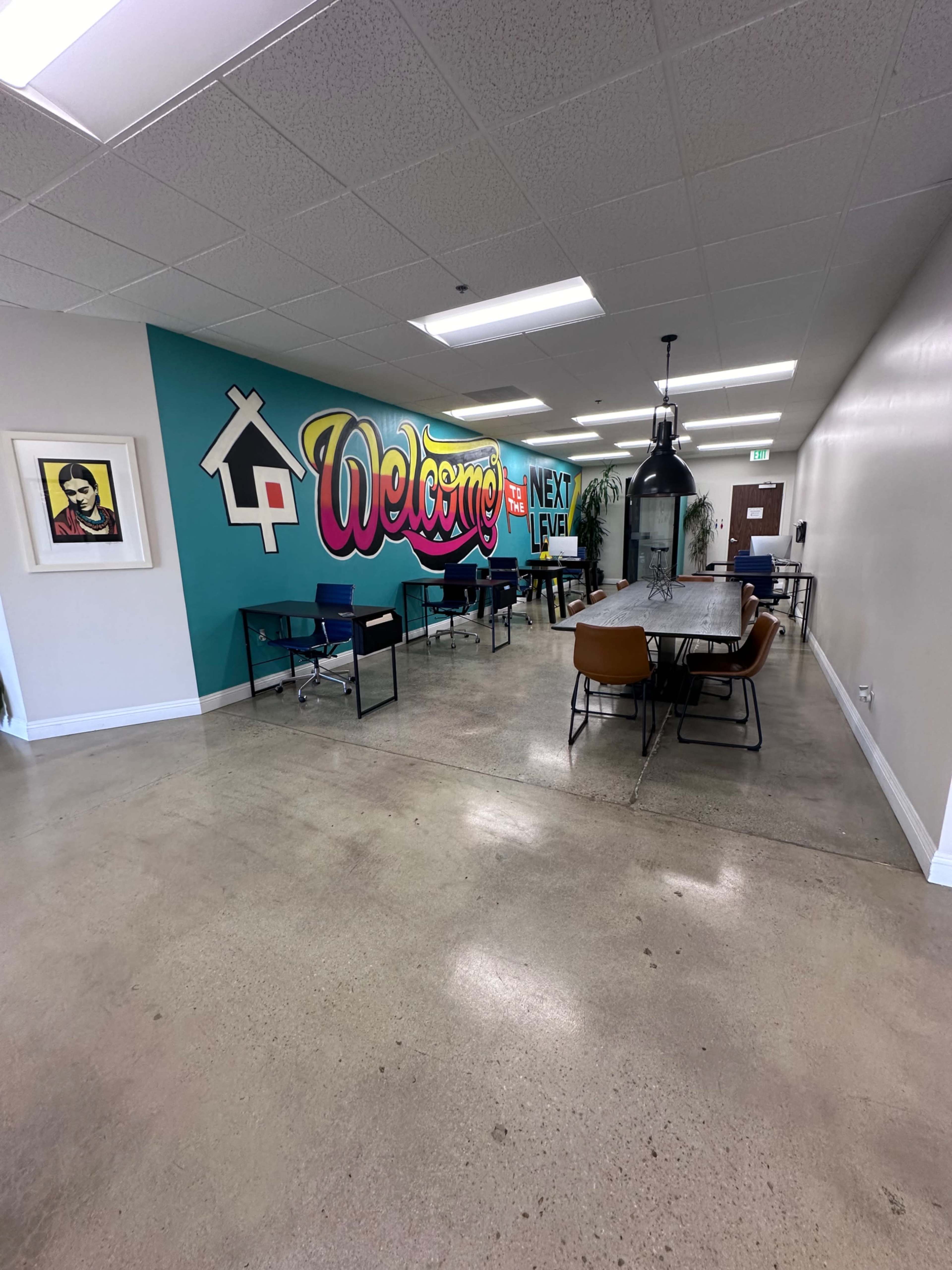 A spacious office area featuring a colorful "Welcome" mural, several desks with chairs, and a large communal table in a well-lit environment.