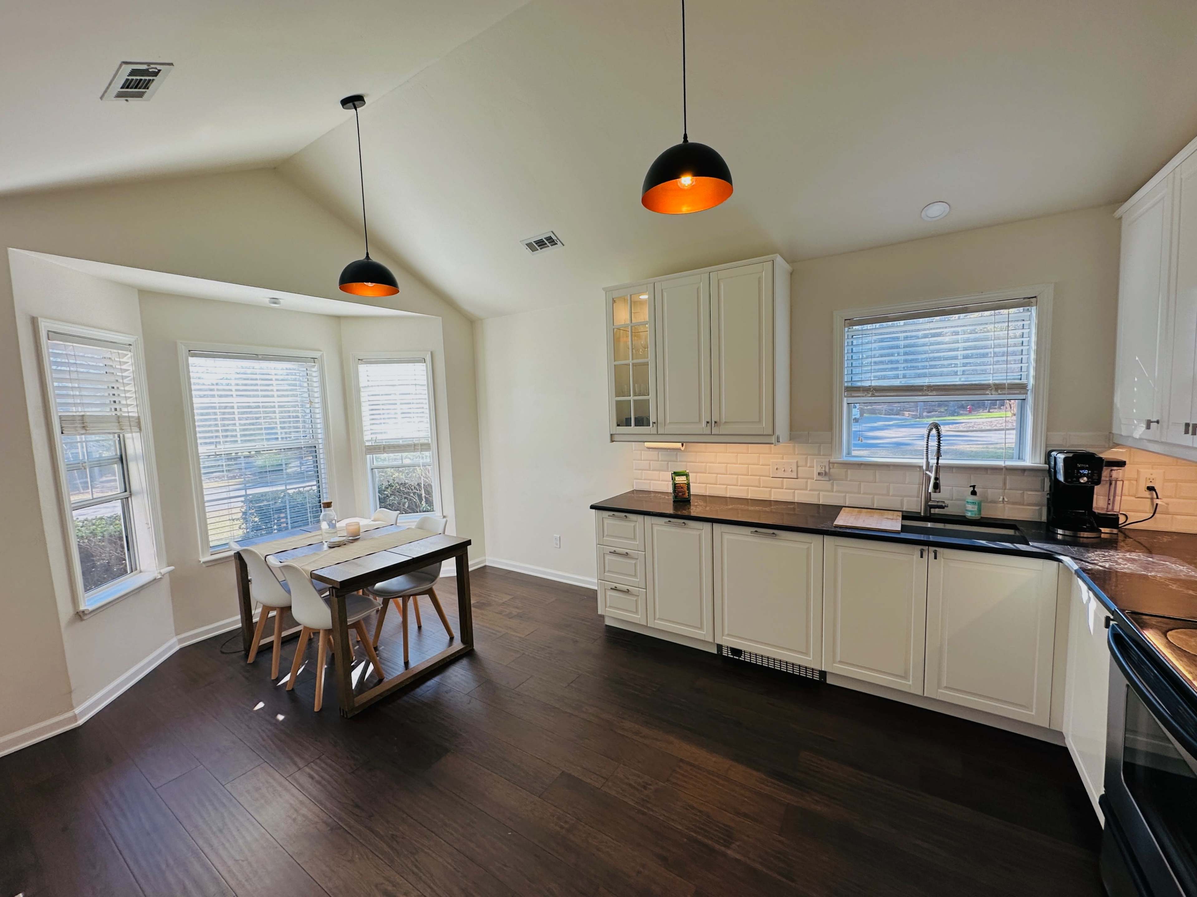 A modern kitchen features white cabinetry, a dark countertop, a dining table with four chairs, and large windows allowing natural light.