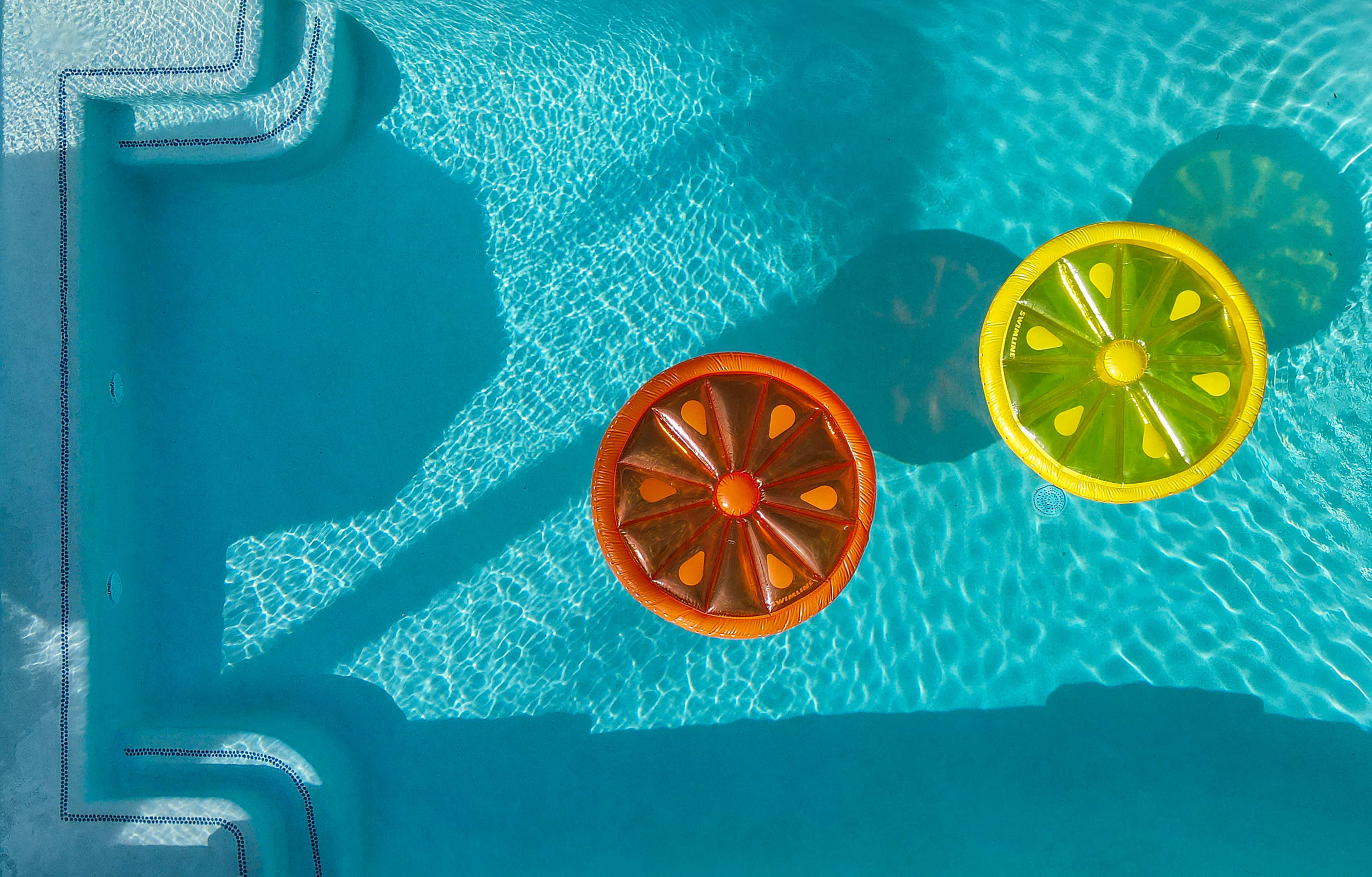 Two round pool floaties shaped like slices of orange and lemon are floating in a clear blue swimming pool.