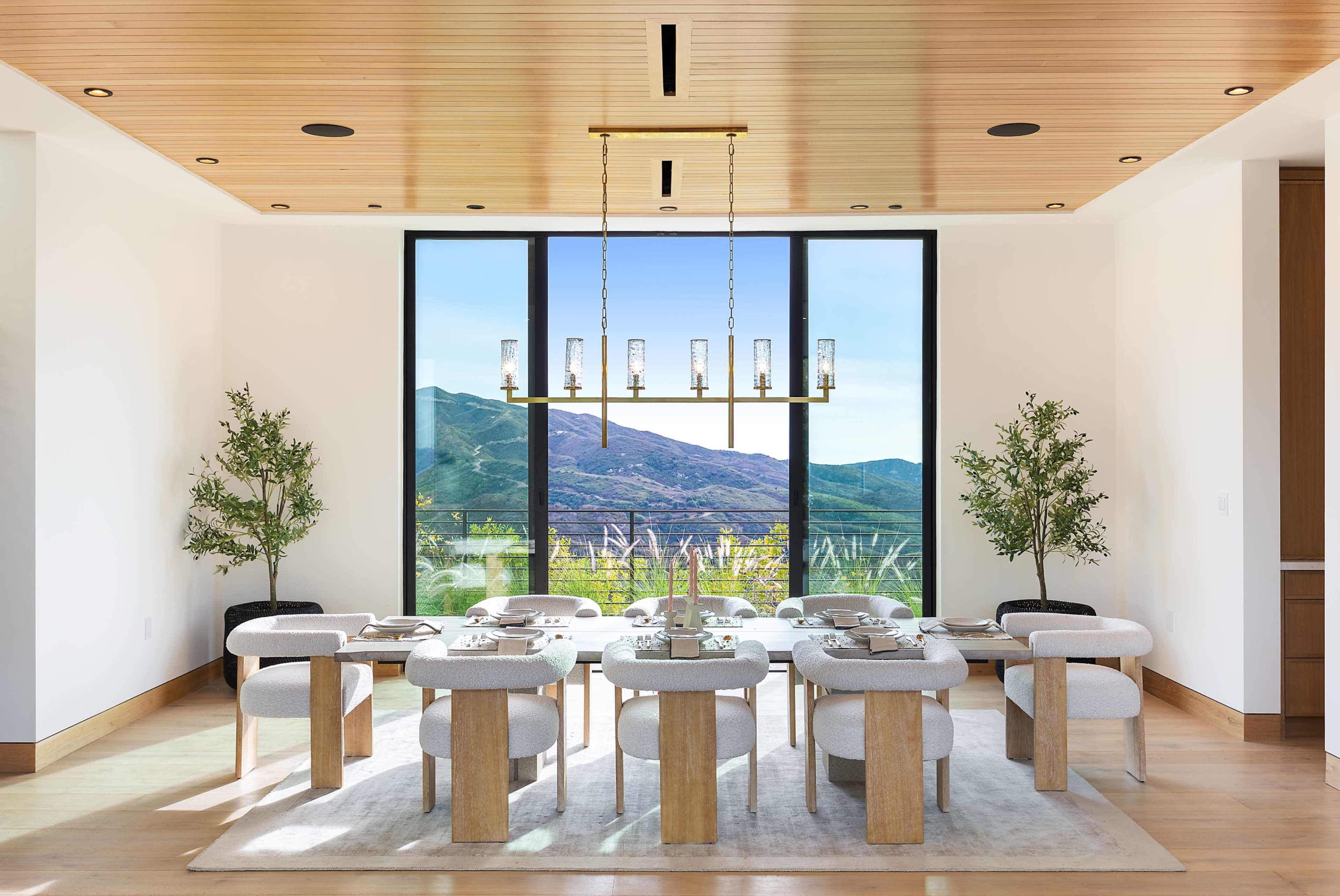 A modern dining room features a long table with plush chairs, surrounded by large windows that overlook a mountainous landscape.