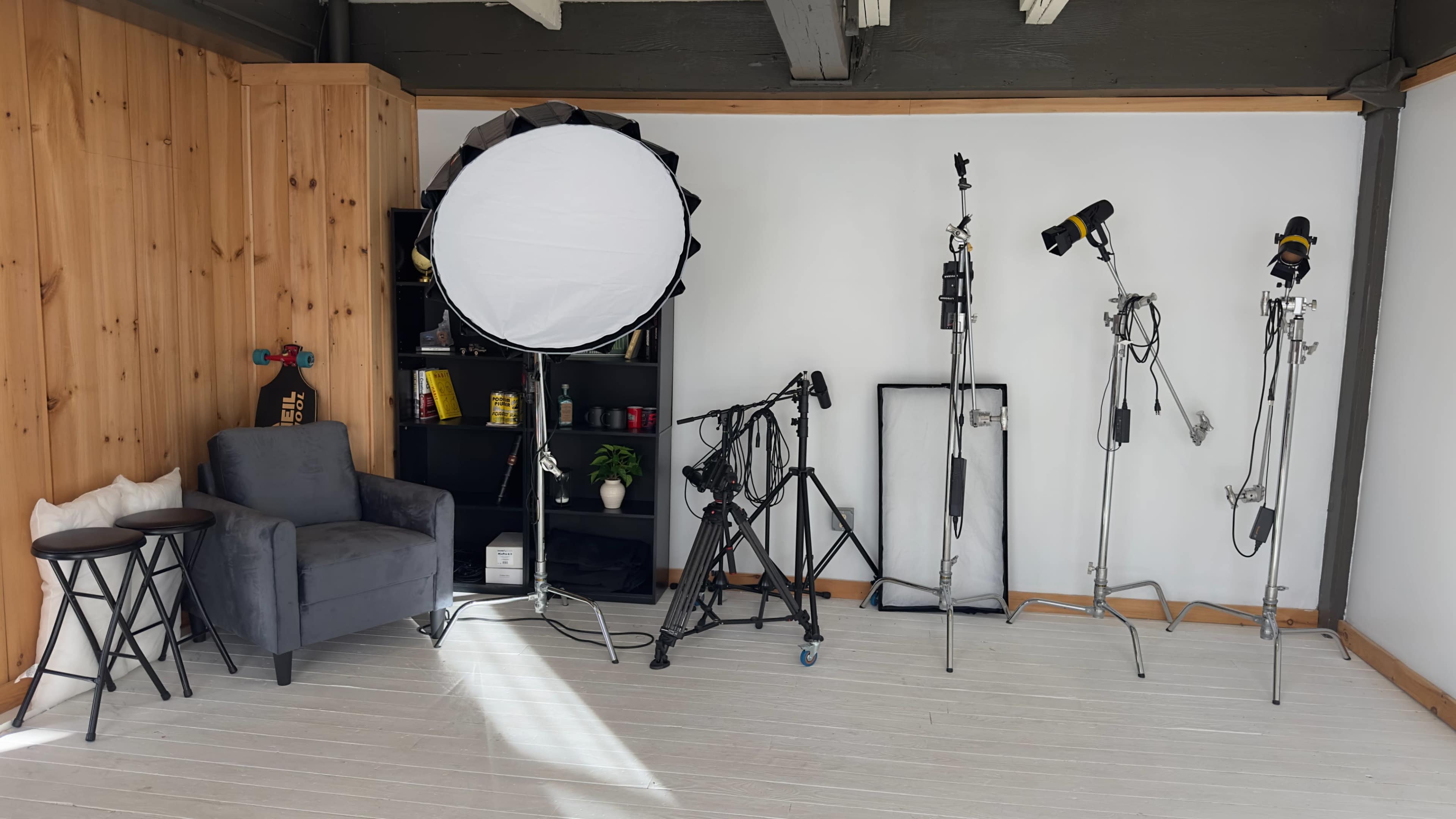 The image shows a photography studio with a grey armchair, a round reflector, various lighting equipment on stands, and a black shelf with props.