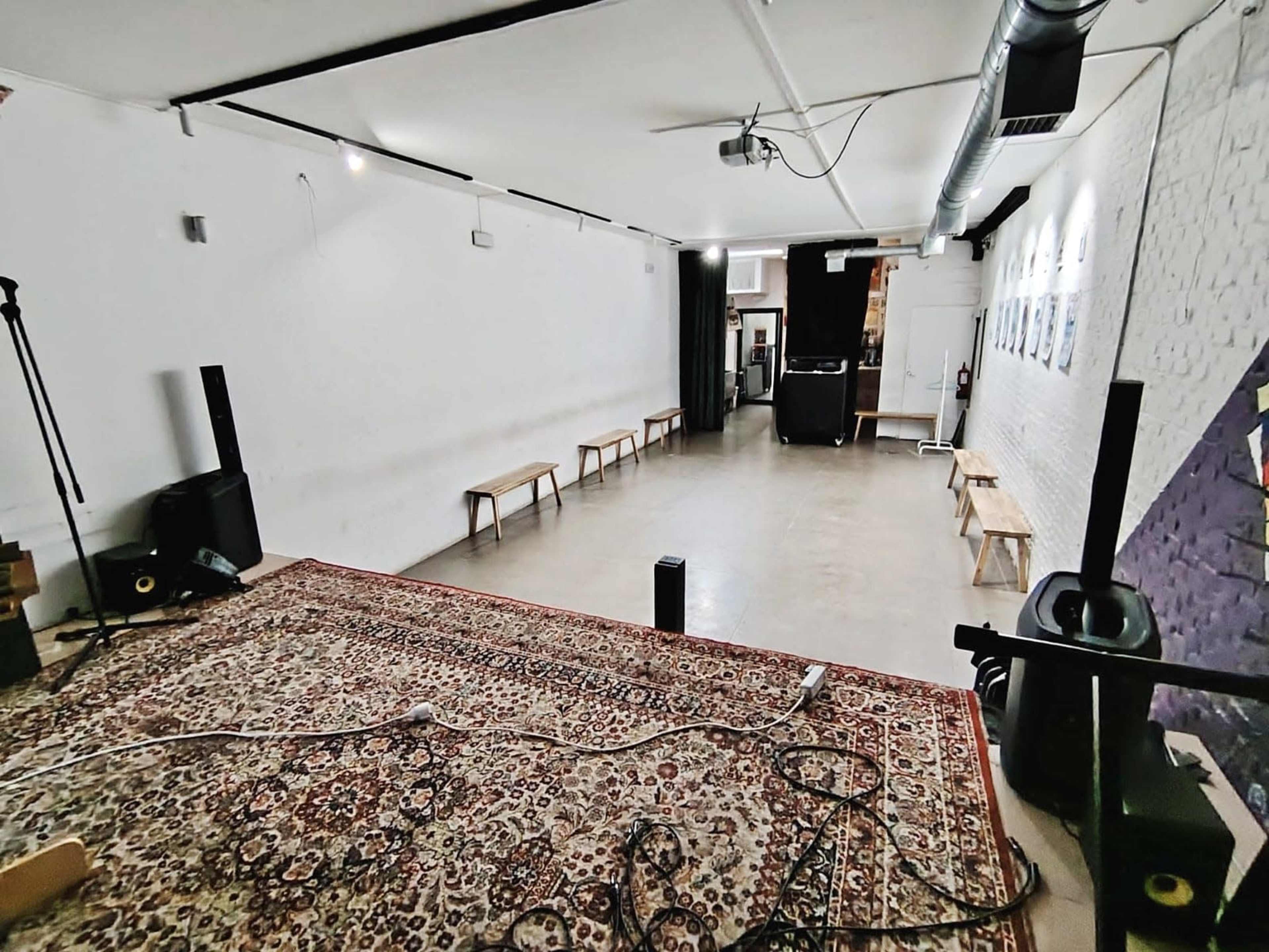 The image shows a spacious, empty performance area with a patterned rug, several benches along the walls, and a sound system setup in a minimalistic room.
