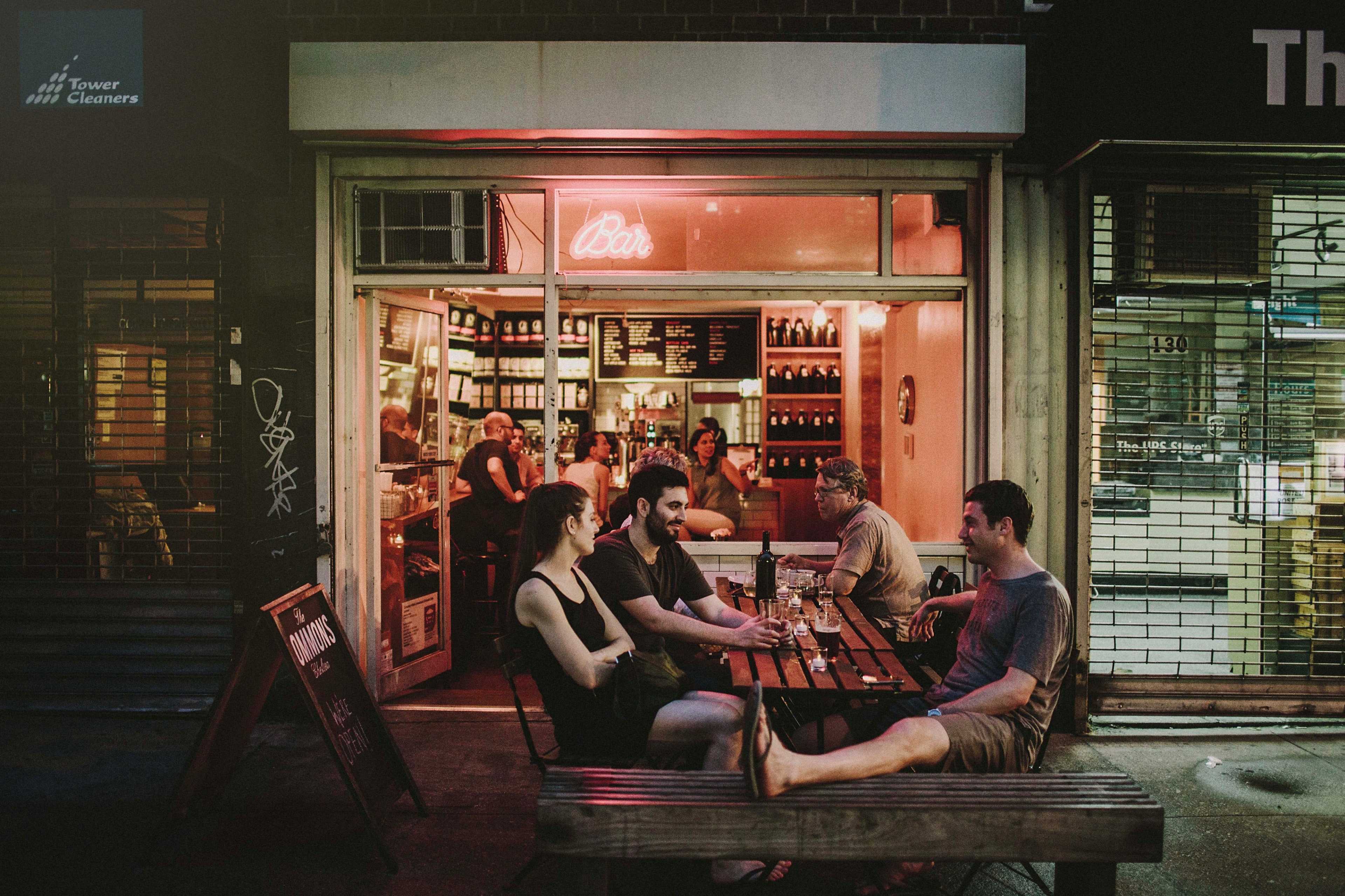 A group of four people sits at an outdoor table in front of a bar, with warm light spilling from inside where other patrons are visible.