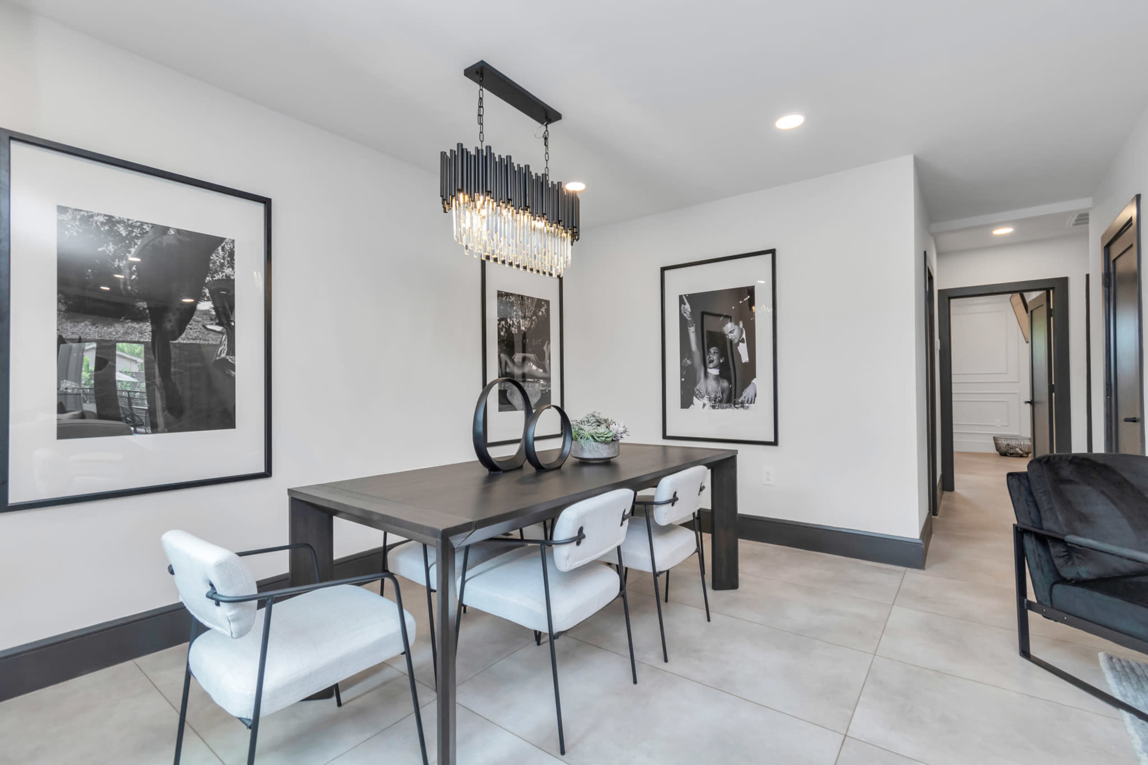 A modern dining area features a rectangular table with white upholstered chairs, framed black-and-white photographs on the walls, and a stylish chandelier overhead.