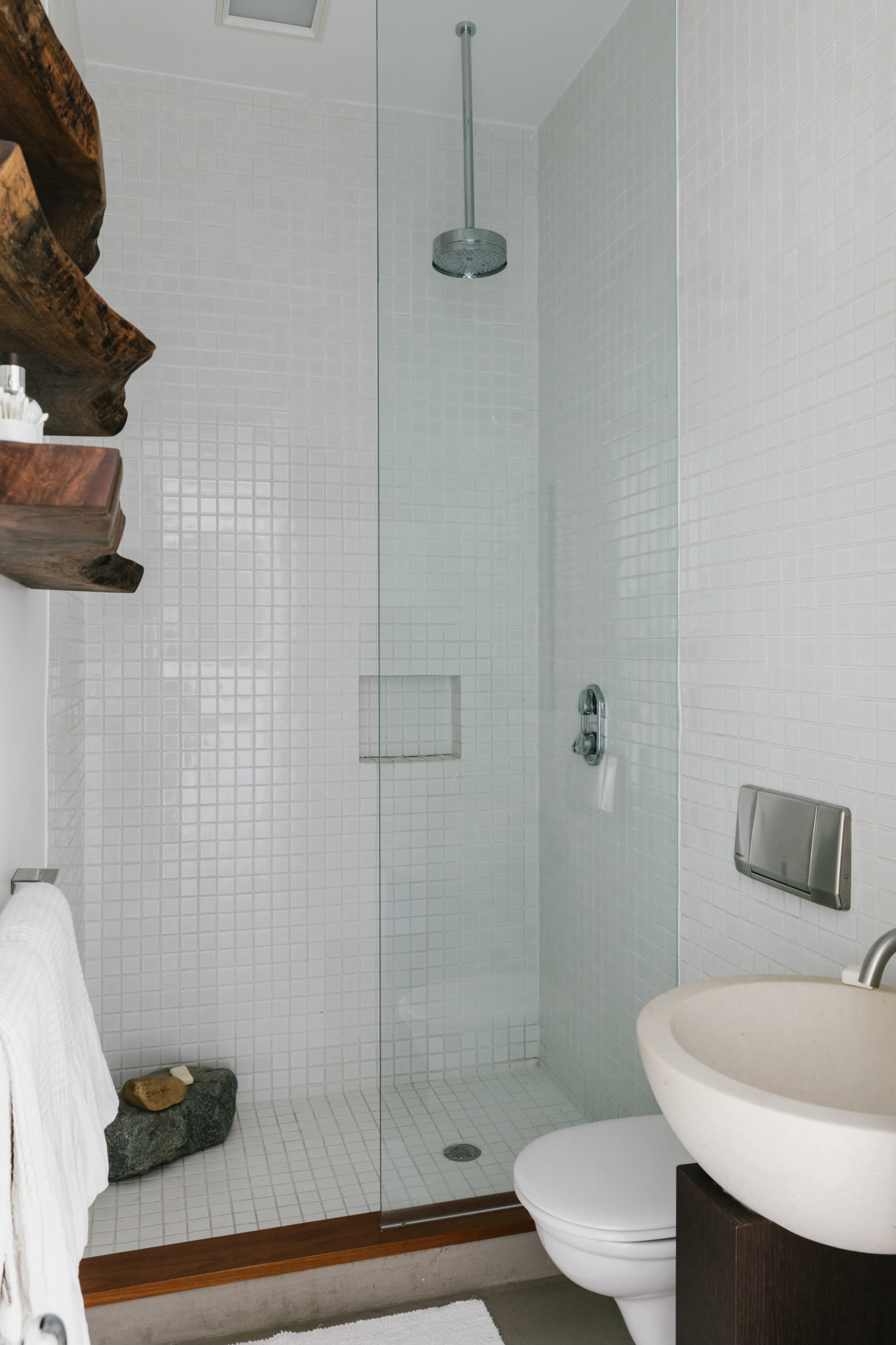 A modern bathroom features a glass shower enclosure, a round sink, and a wooden shelf, all with white tiled walls.