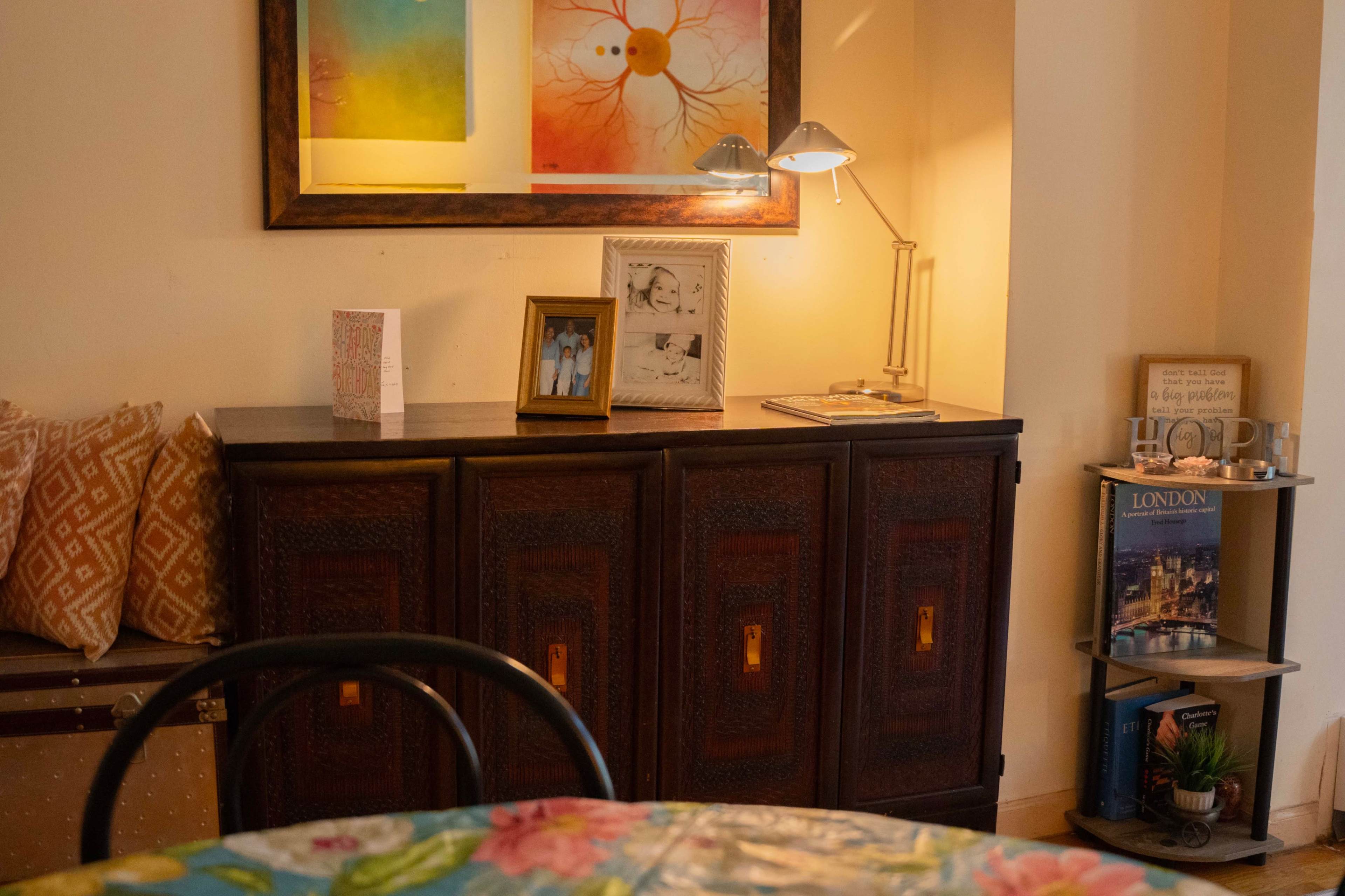 The image shows a cozy interior with a wooden cabinet, framed photos, a modern lamp, and a small bookshelf displaying books and decorative items.