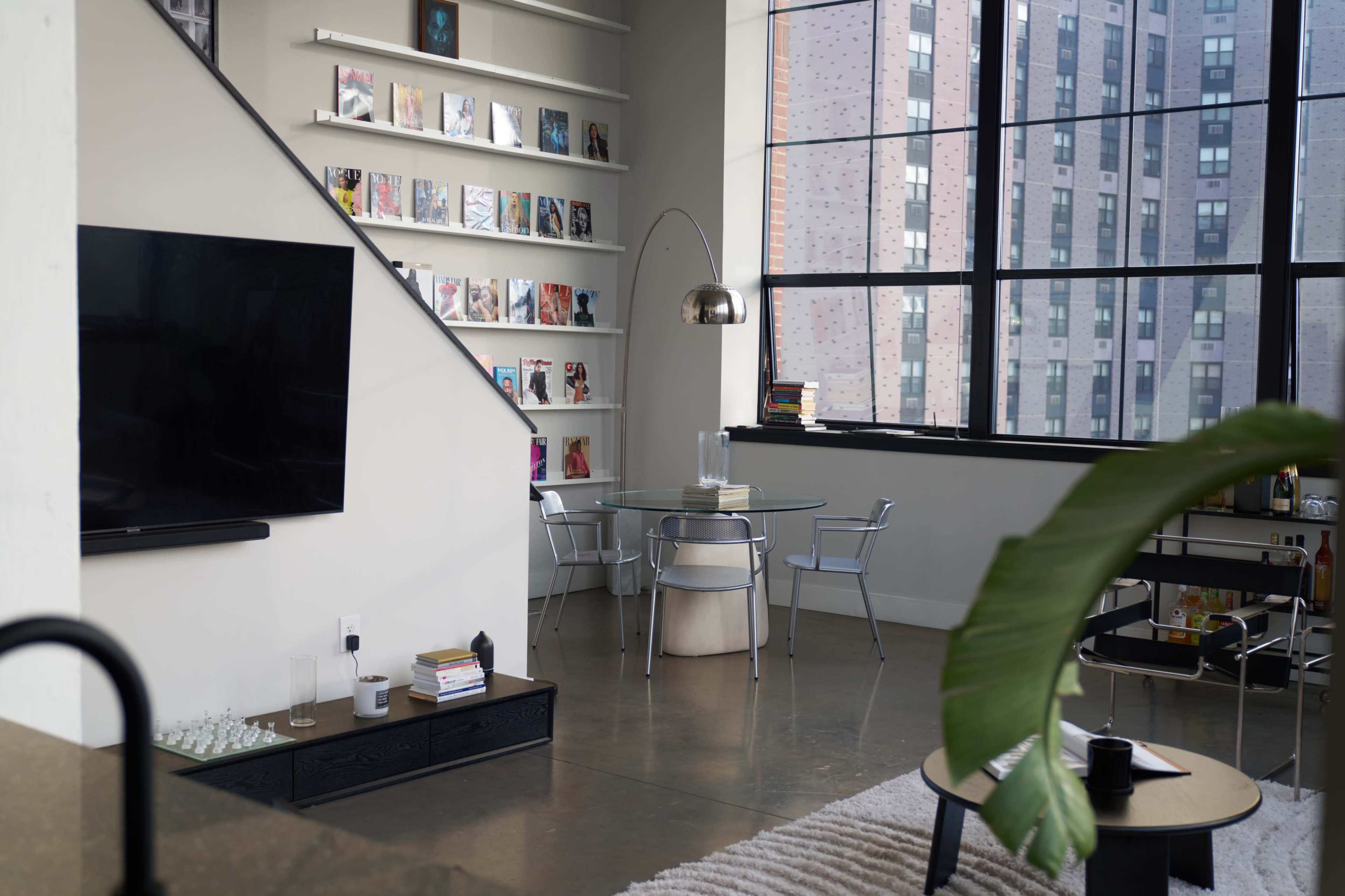 A modern living area featuring a large window, a television mounted on a wall, a glass dining table with steel chairs, and a bookshelf displaying magazines.
