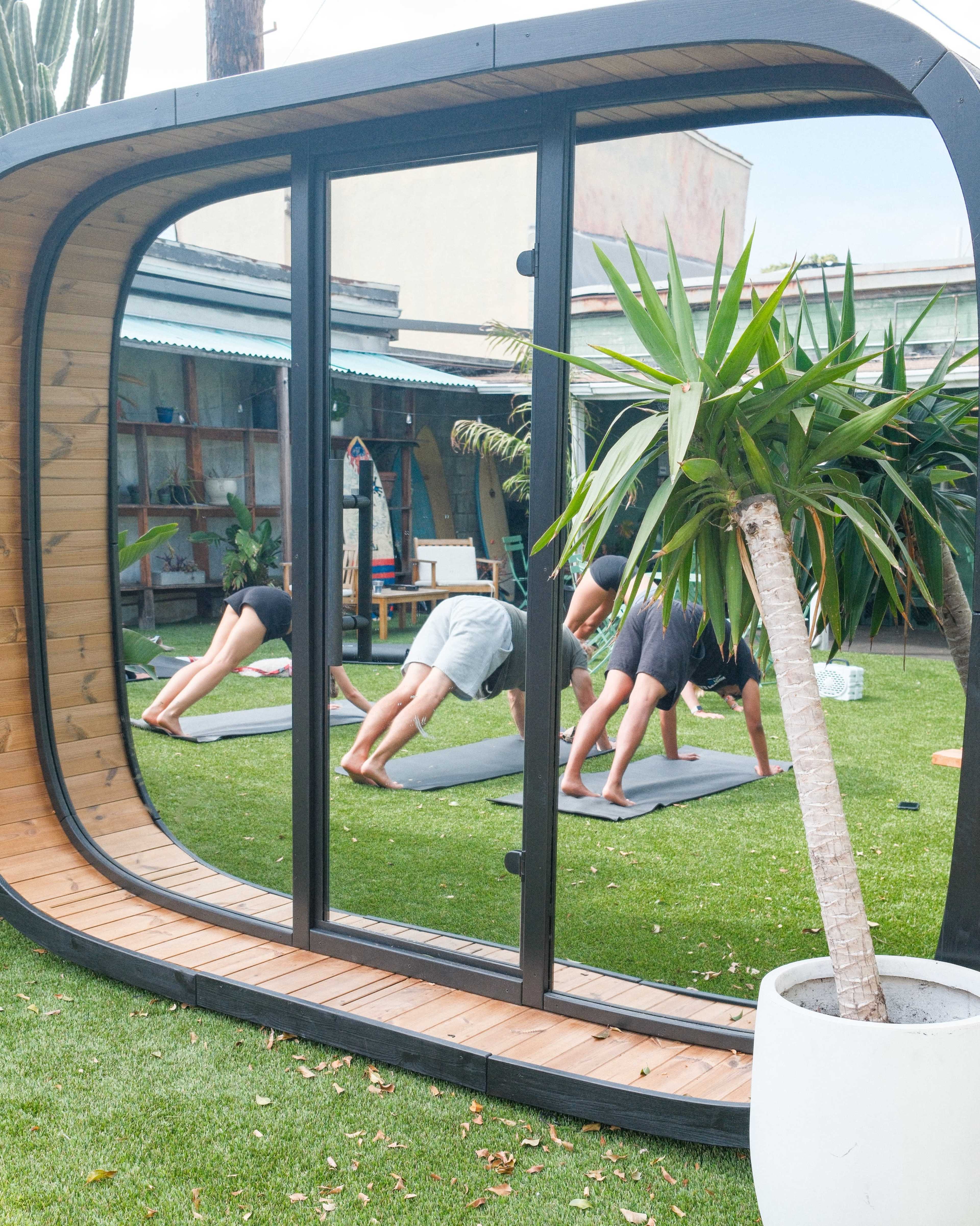 A group of people practices yoga in a lush outdoor space, framed by a modern curved structure with glass walls.