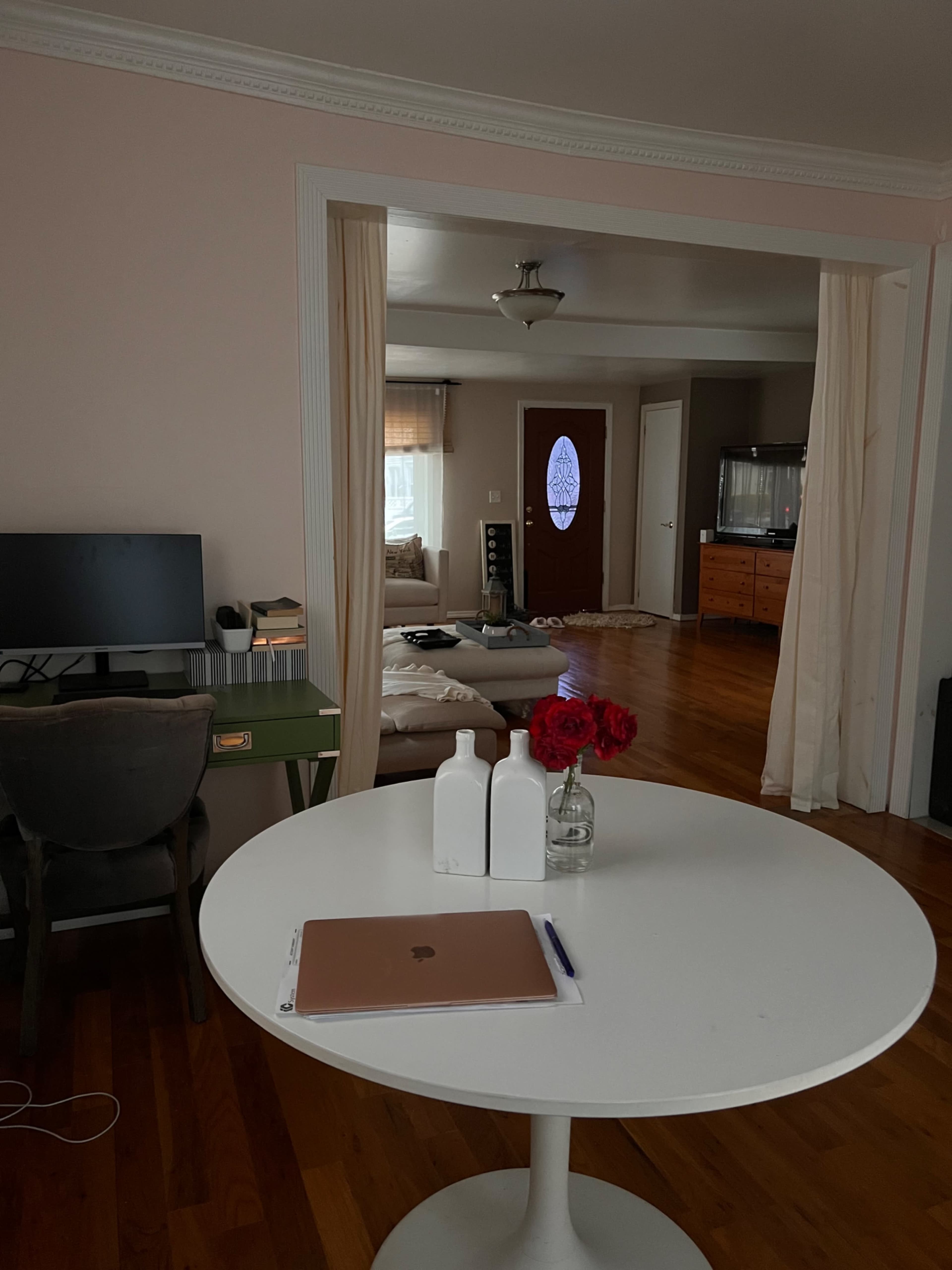 A modern living space features a round white table with a laptop and decorative items, viewed from an adjacent room with a glimpse of furniture and an entrance.