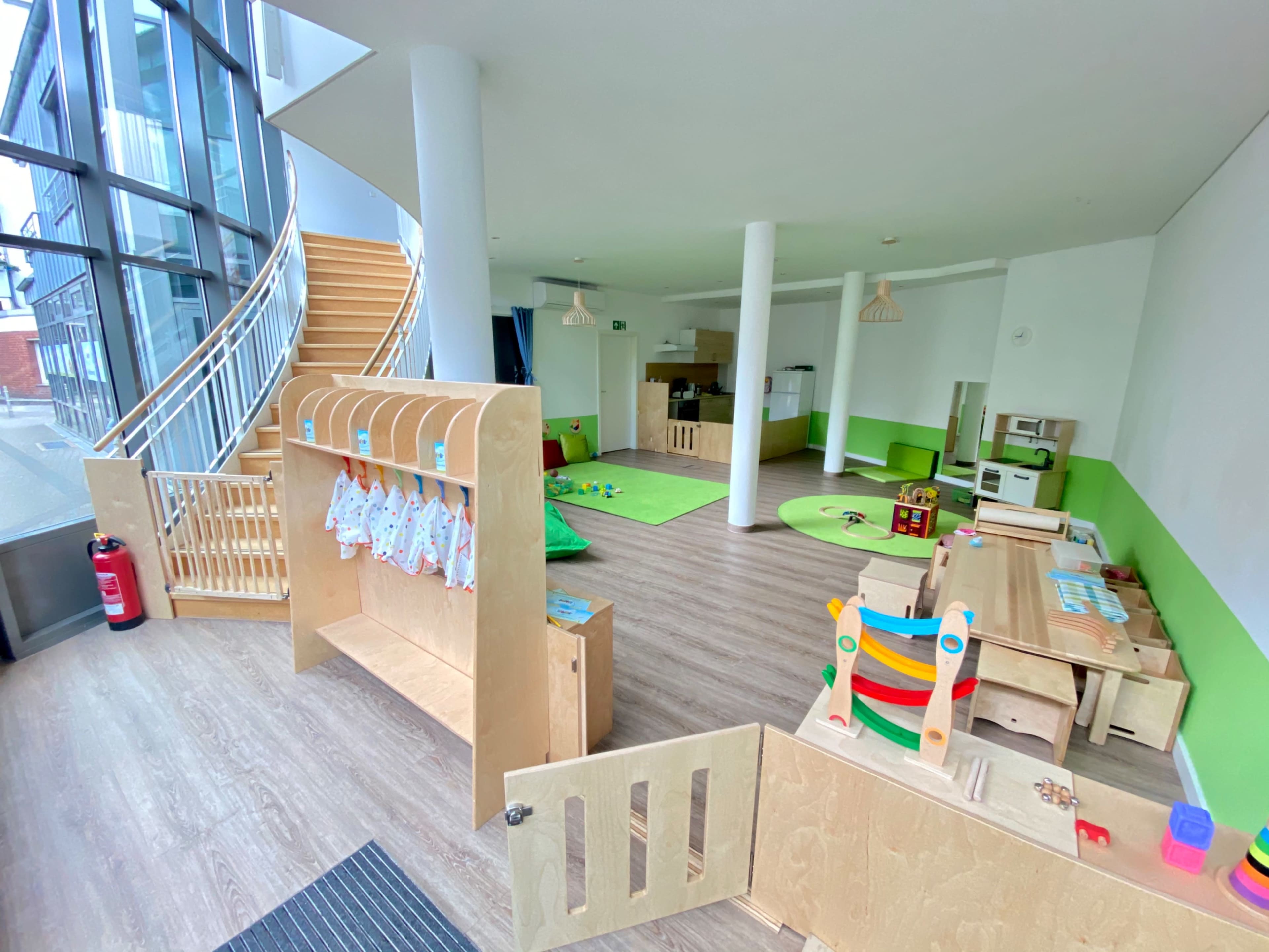The image shows a brightly lit indoor play area for children, featuring a variety of toys, a green play mat, and a staircase in the background.