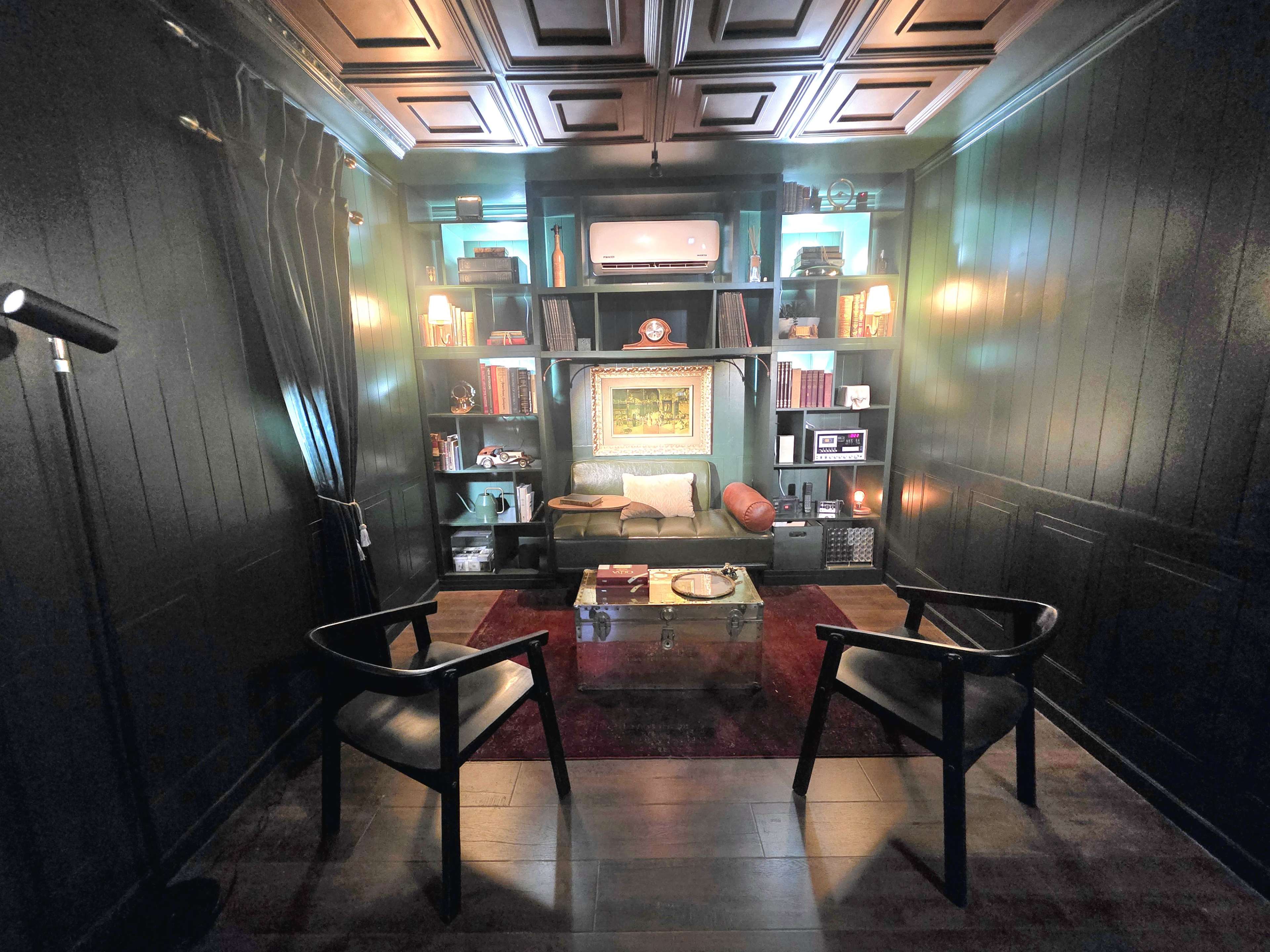The image shows a cozy, dark-paneled room with a pair of black chairs facing a glass coffee table, surrounded by bookshelves filled with books and decorative items.