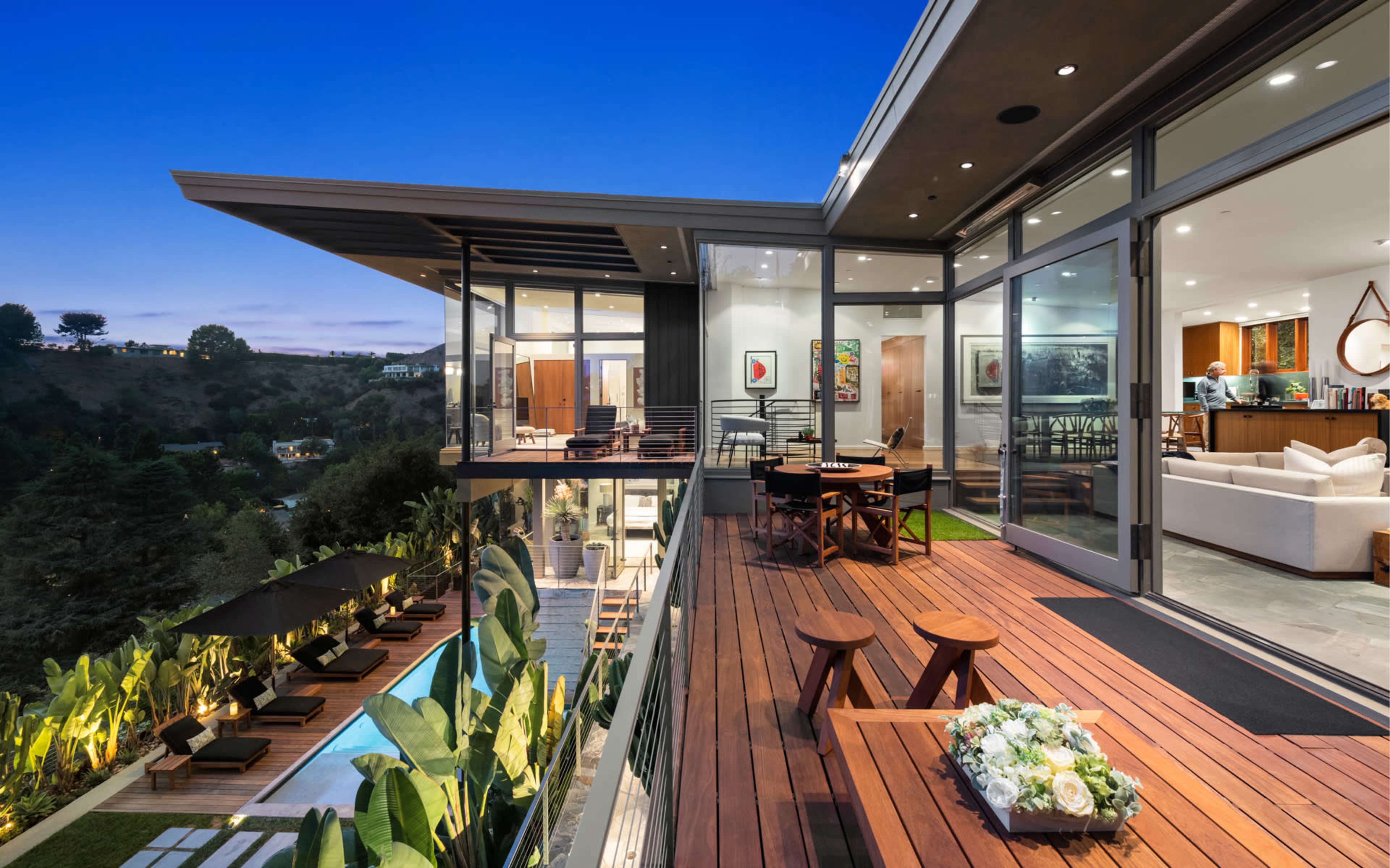 The image shows a modern two-story house with large glass walls, a wooden deck, an outdoor seating area, and a swimming pool surrounded by greenery.