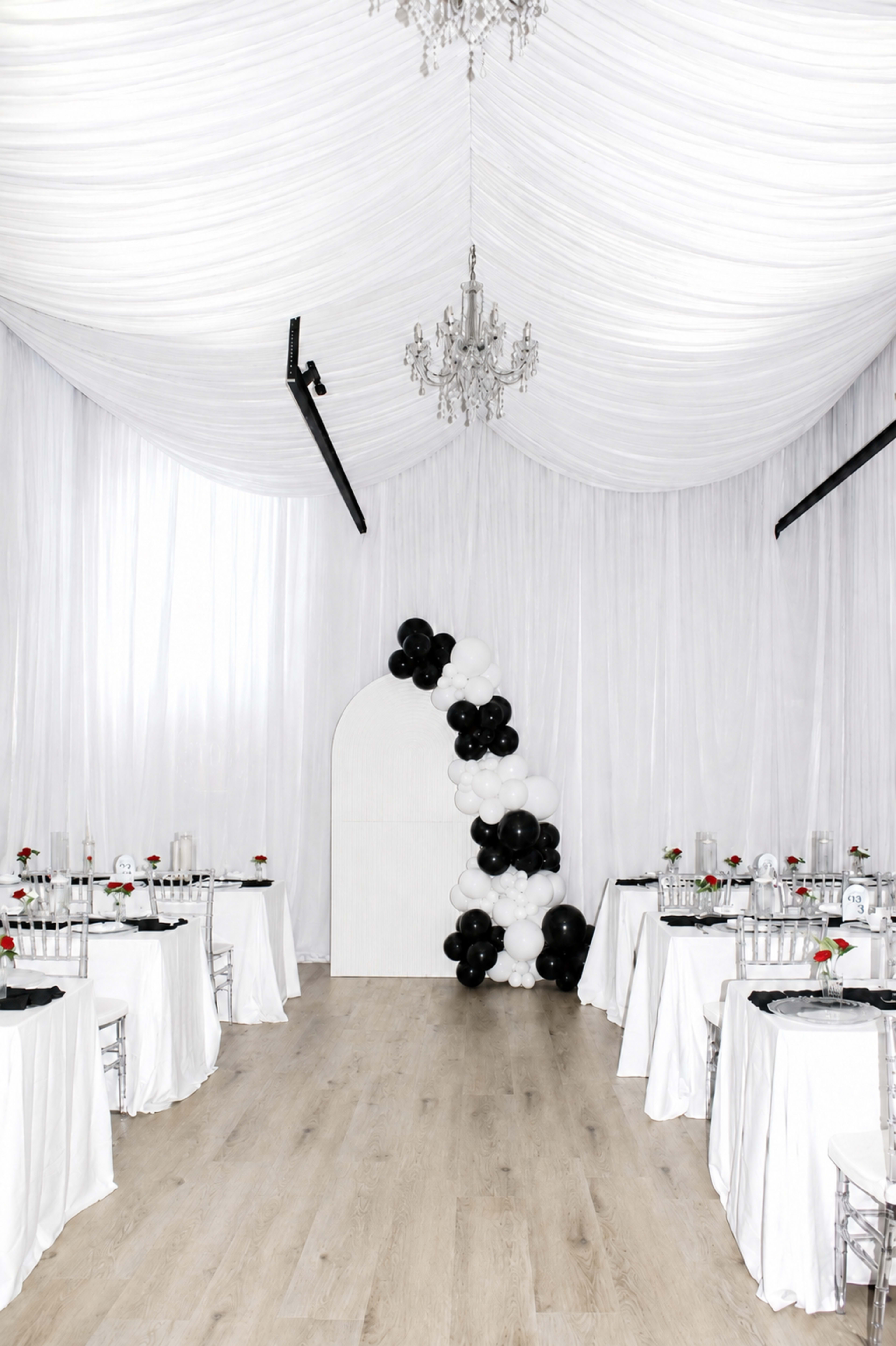A banquet hall features white draped ceilings, black and white balloon decor, and elegantly set tables with floral centerpieces.