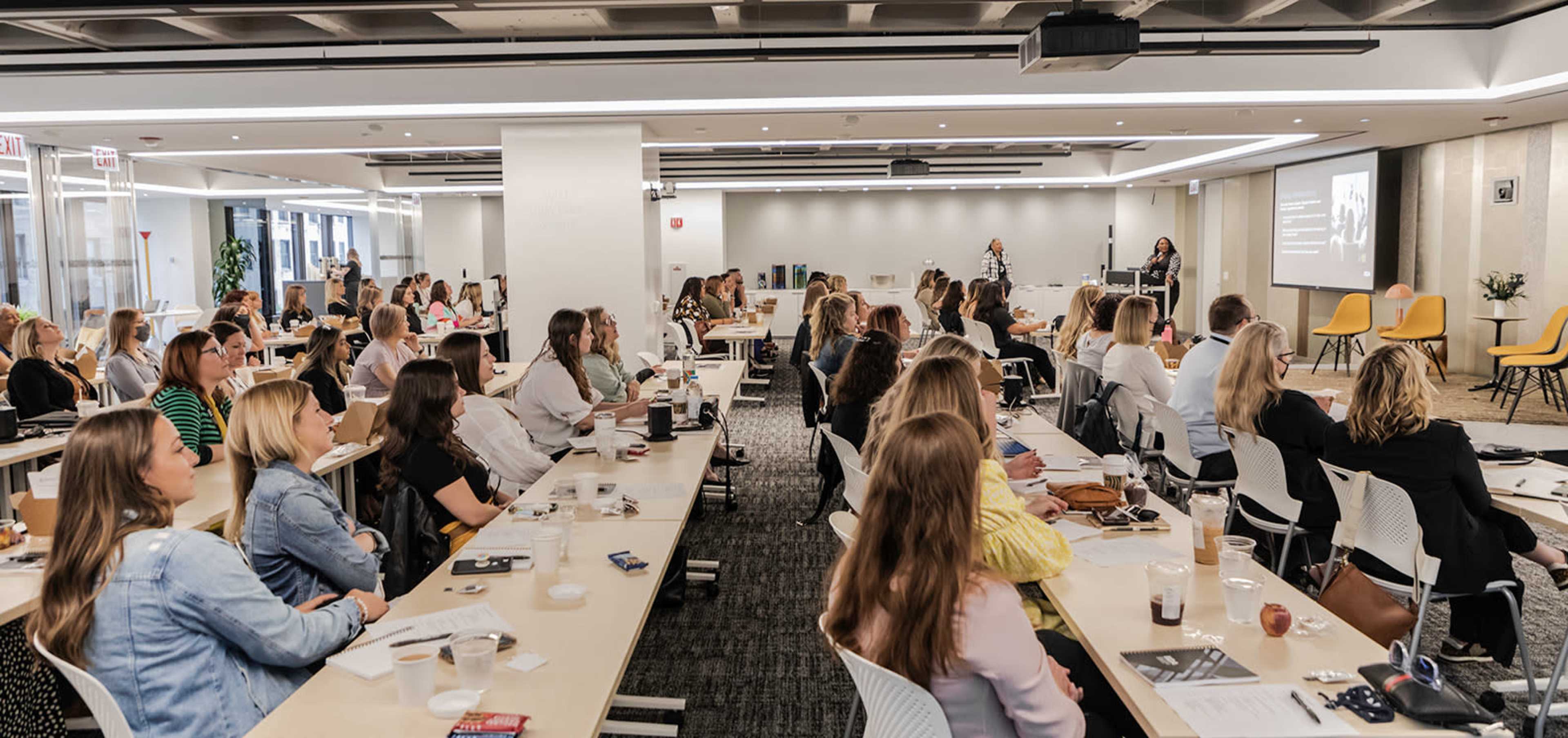 A large group of women is seated in a modern conference room, attentively listening to a speaker at the front of the room.