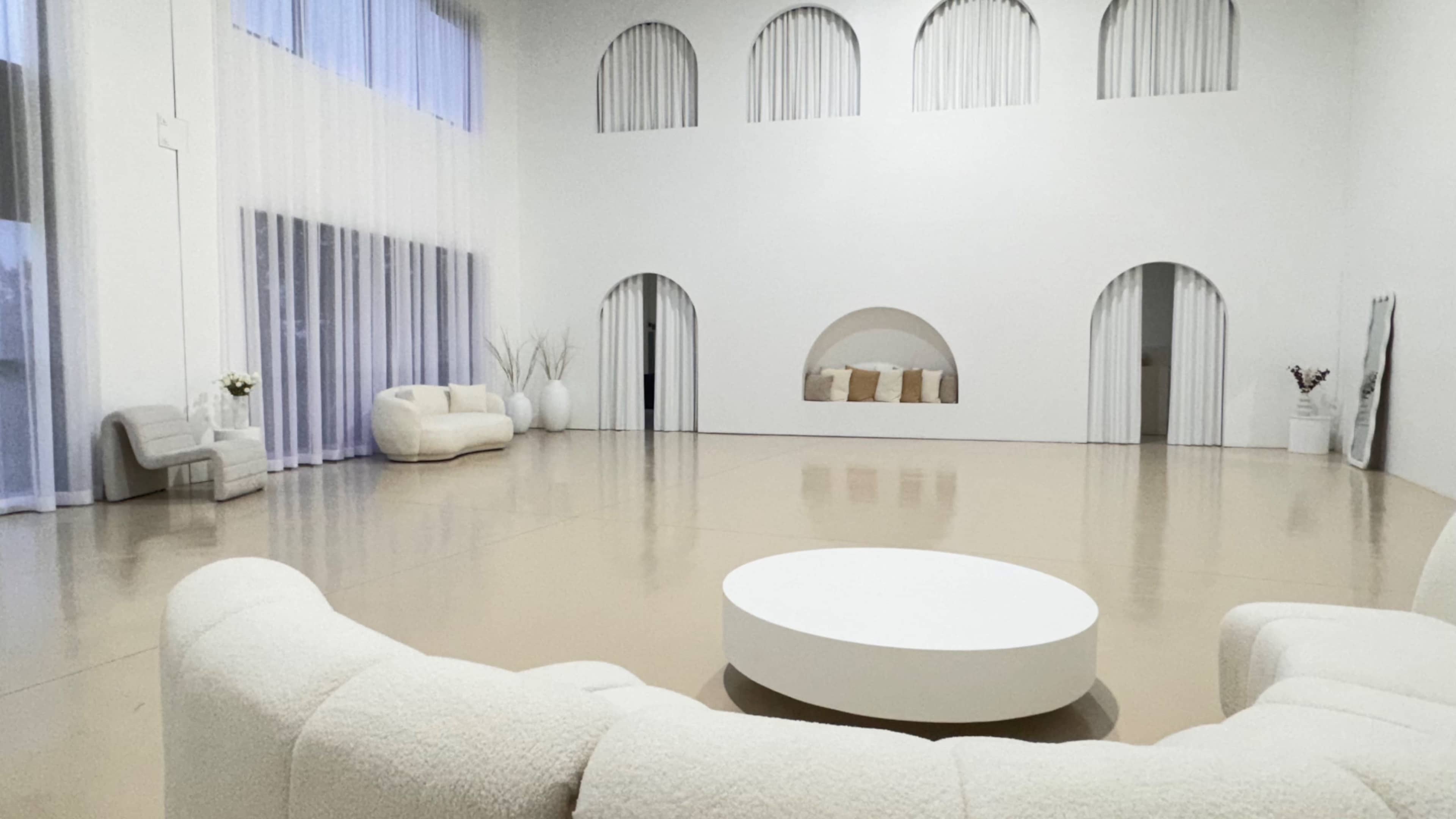 The image shows a spacious, modern interior with a light-colored floor, curved architecture, and minimalist furniture arrangements.