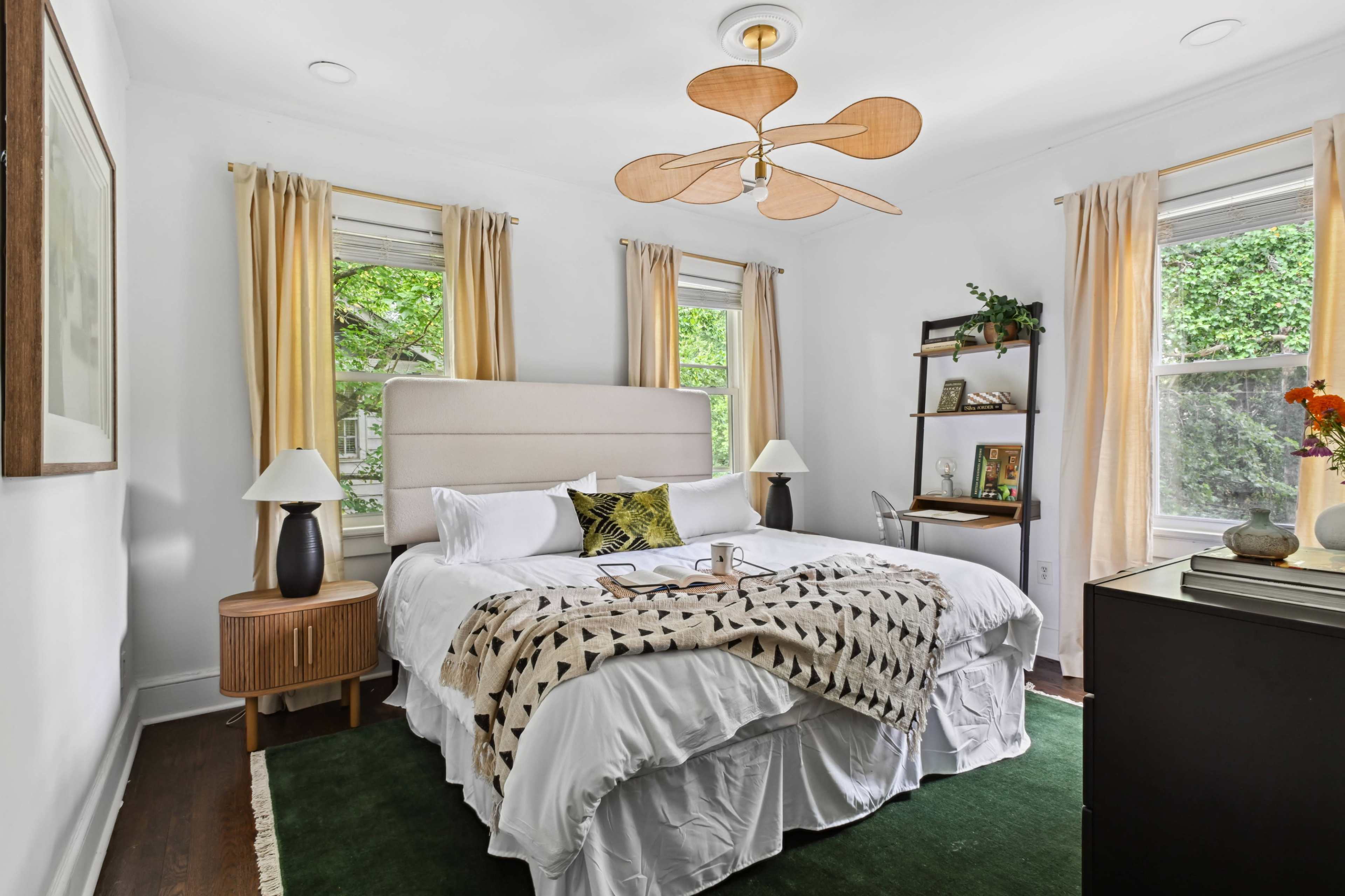 A neatly arranged bedroom with a large bed, two windows adorned with yellow curtains, and a ceiling fan overhead.
