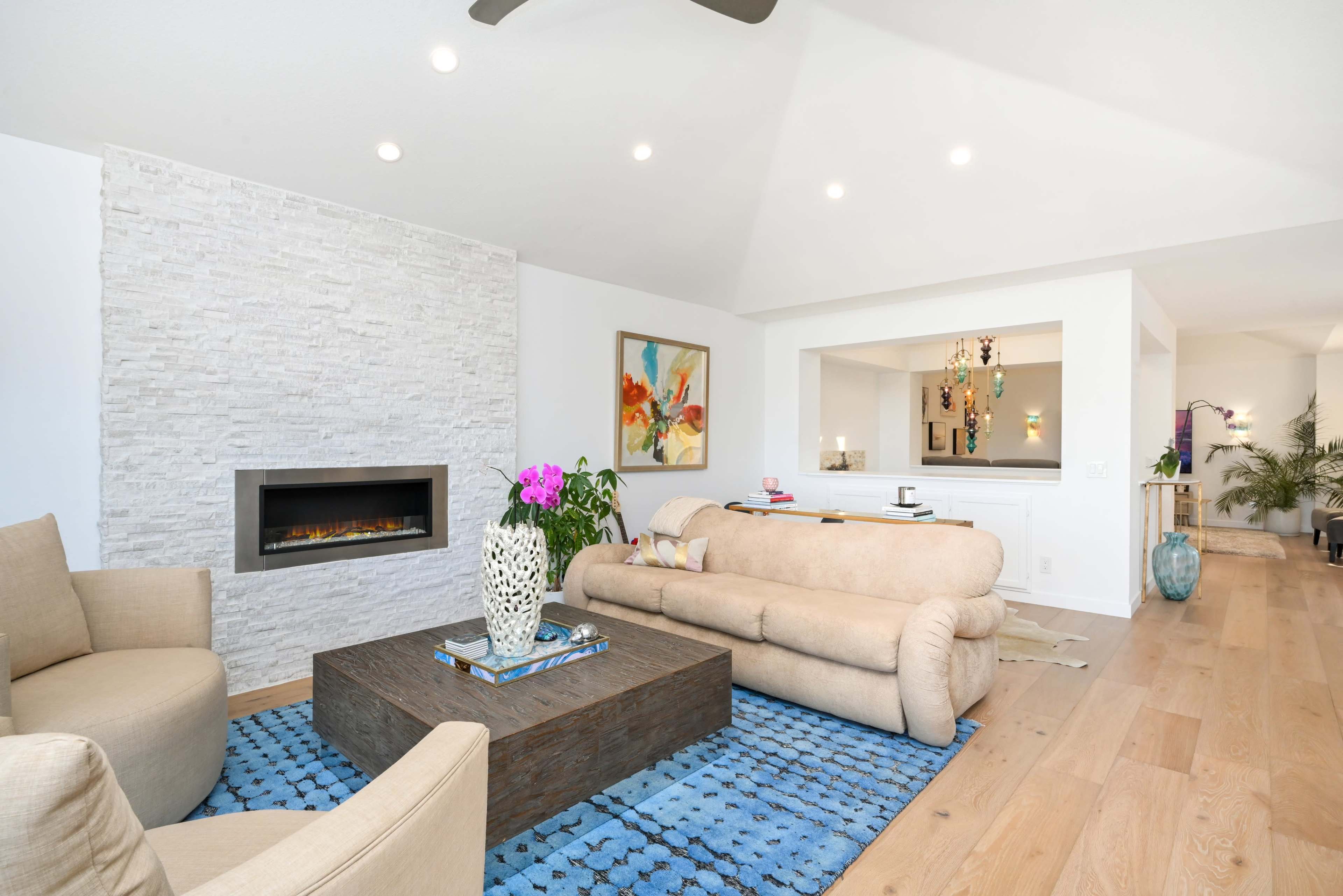 A modern living room featuring a beige sofa, two armchairs, a coffee table, and a fireplace, with colorful artwork and decorative plants in the background.