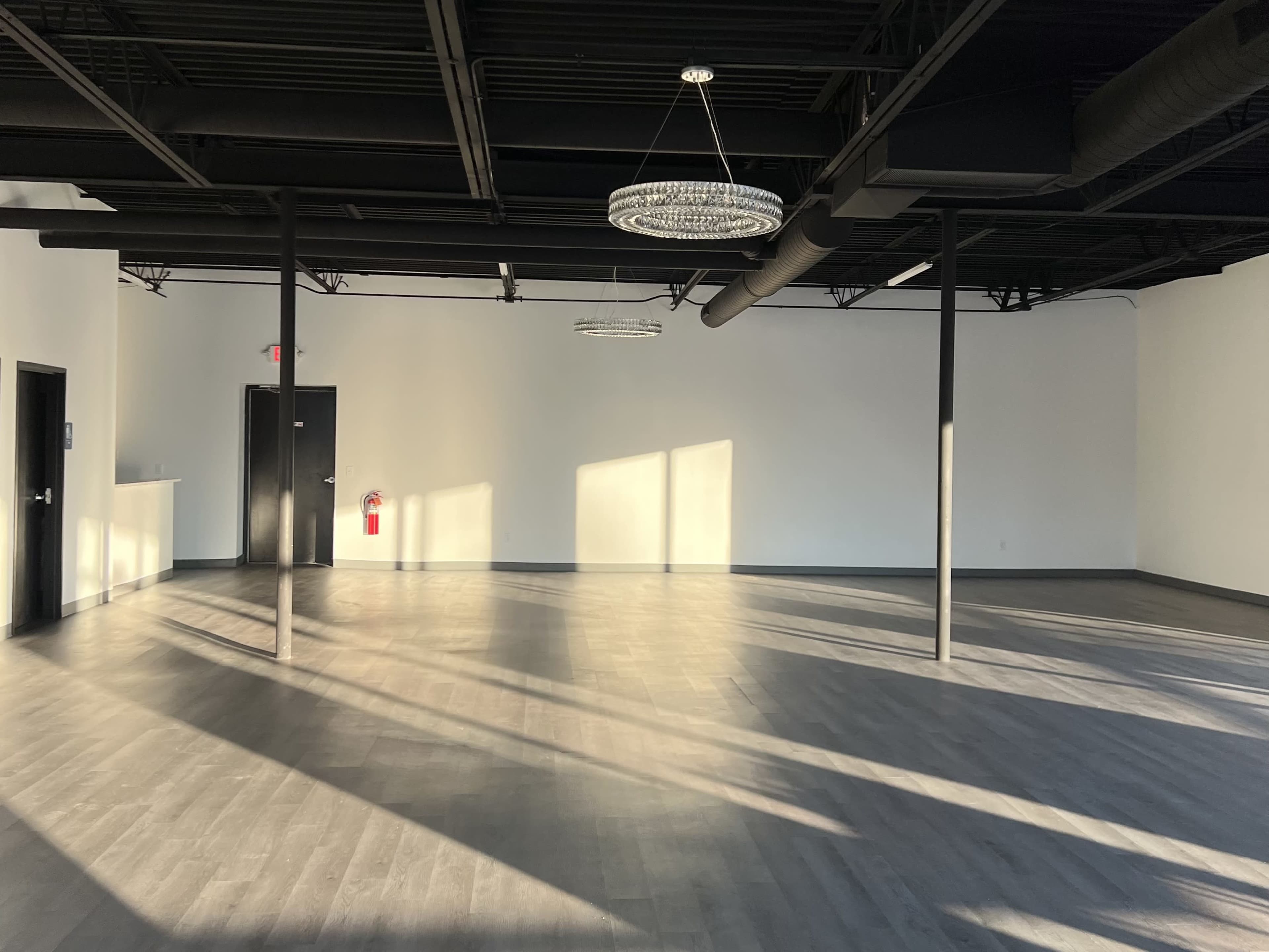 The image depicts a spacious, empty room with wooden flooring, illuminated by sunlight casting long shadows from a pair of modern light fixtures.