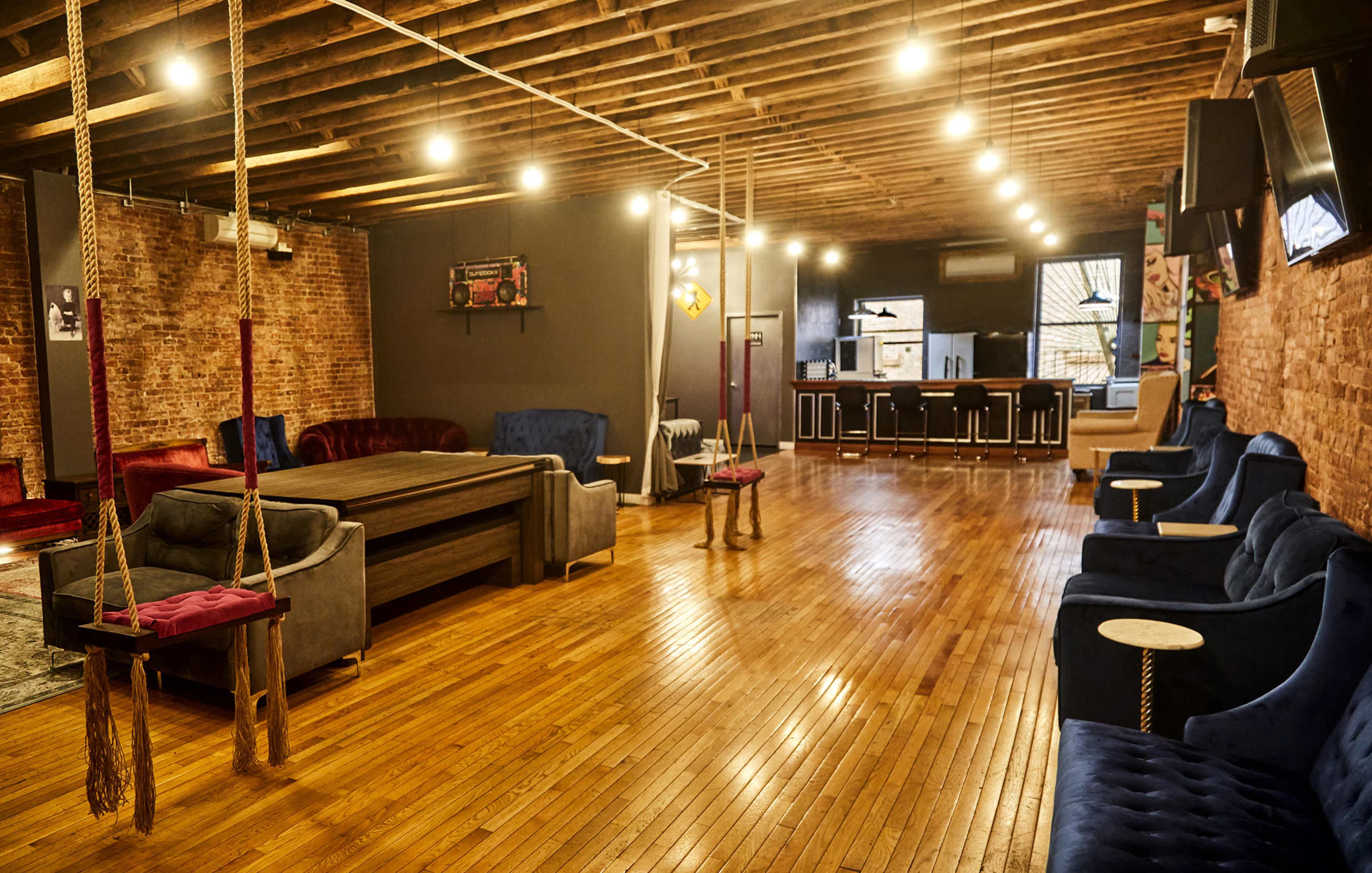 A spacious indoor area with wooden flooring, featuring various seating arrangements, including sofas and swings, and a bar counter at the far end.