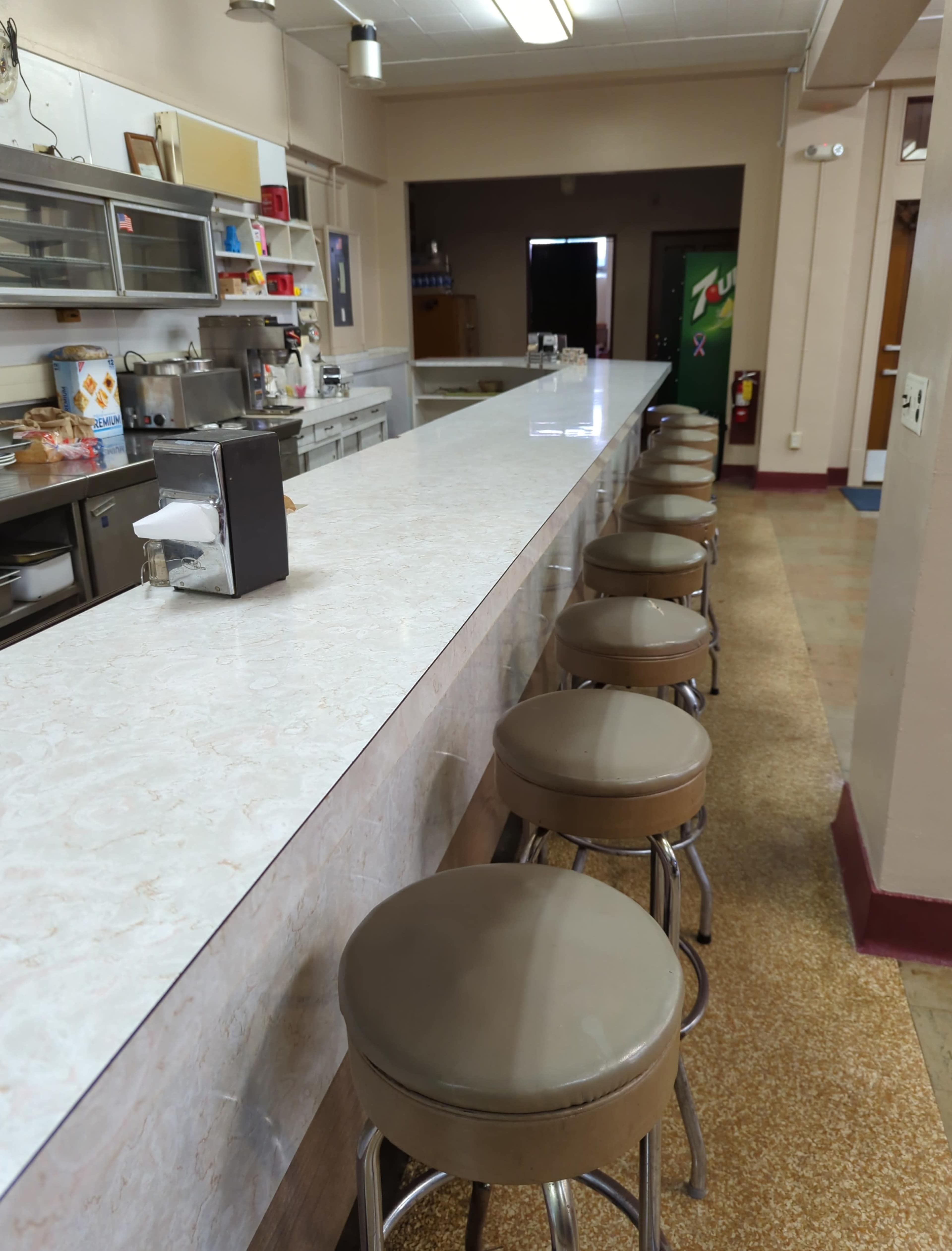 A clean diner-style counter with several brown stools line one side, while the other side features a kitchen area.